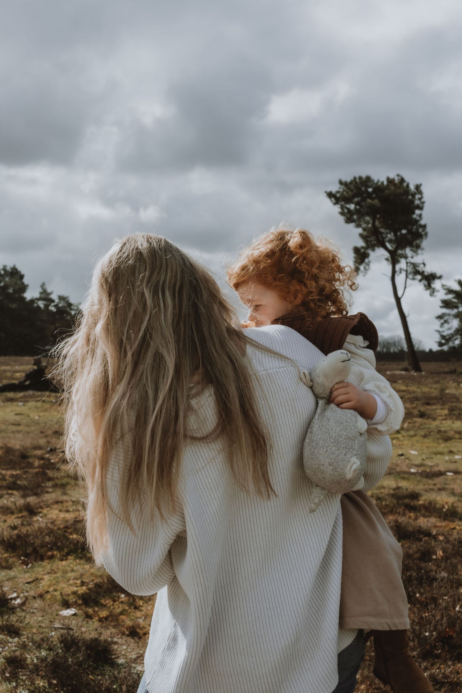 Blonde mother carrying curly redhead toddler in open heathland under overcast sky.