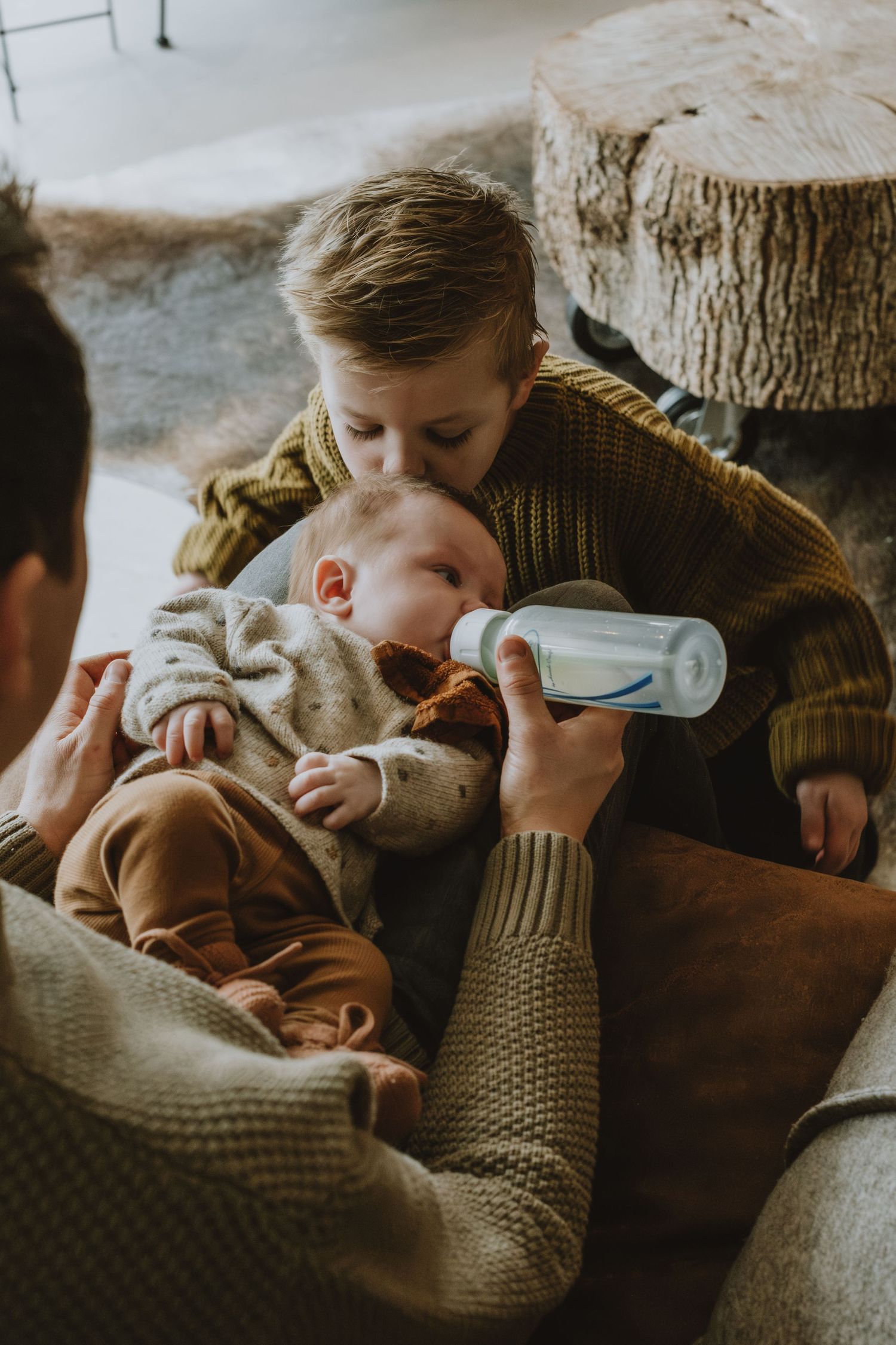 Young boy kissing newborn sibling while bottle feeding in cozy home setting.