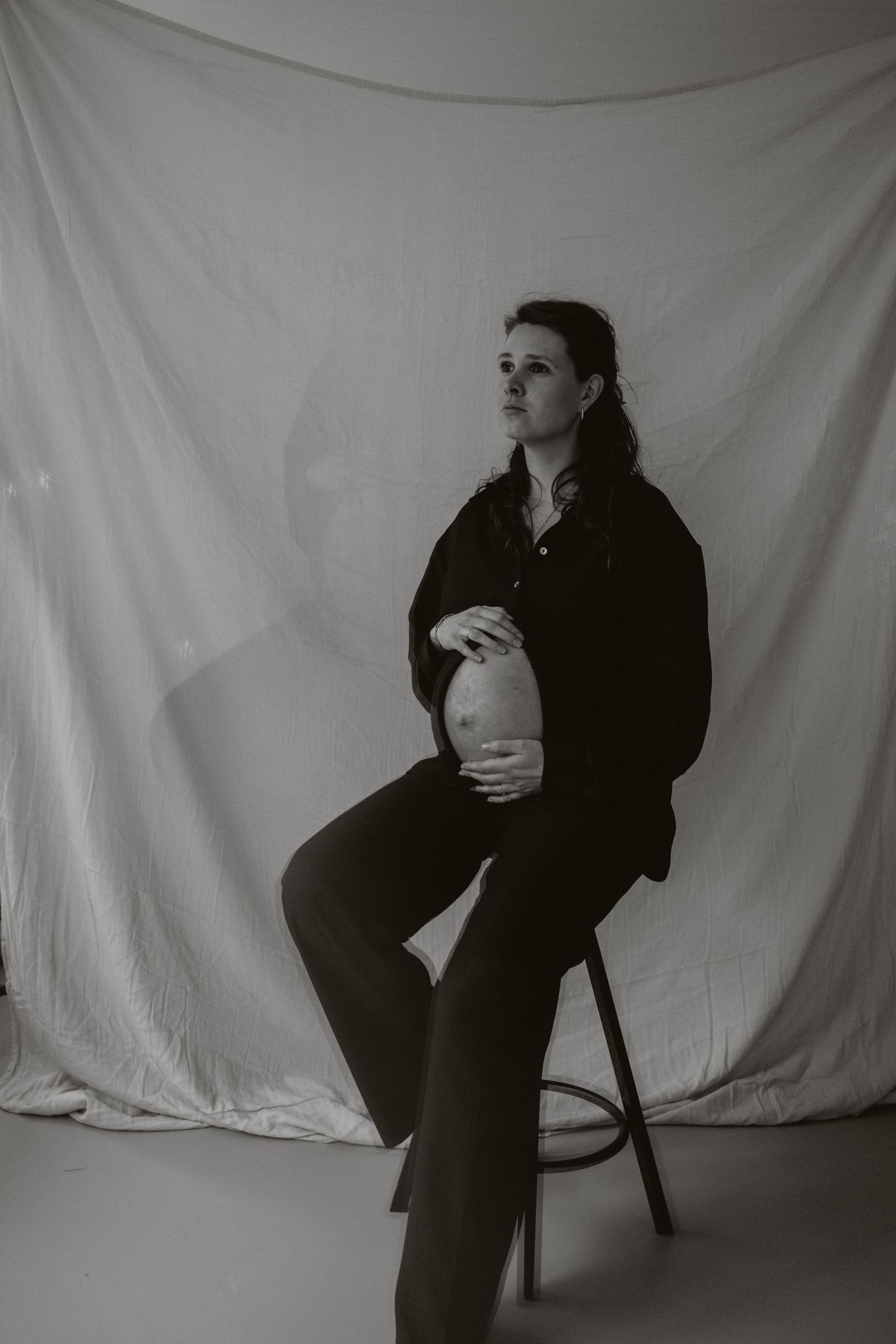 Black and white maternity portrait of woman sitting on stool holding belly before white backdrop.
