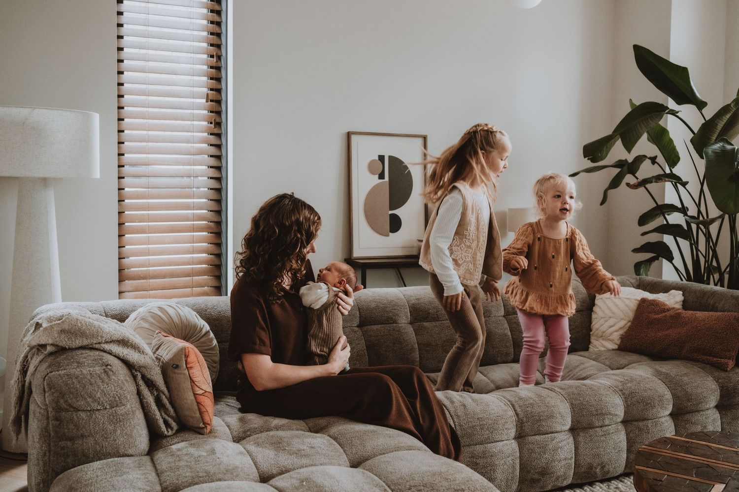 Mother sits on sofa holding newborn while two young daughters play nearby in bright modern living room.