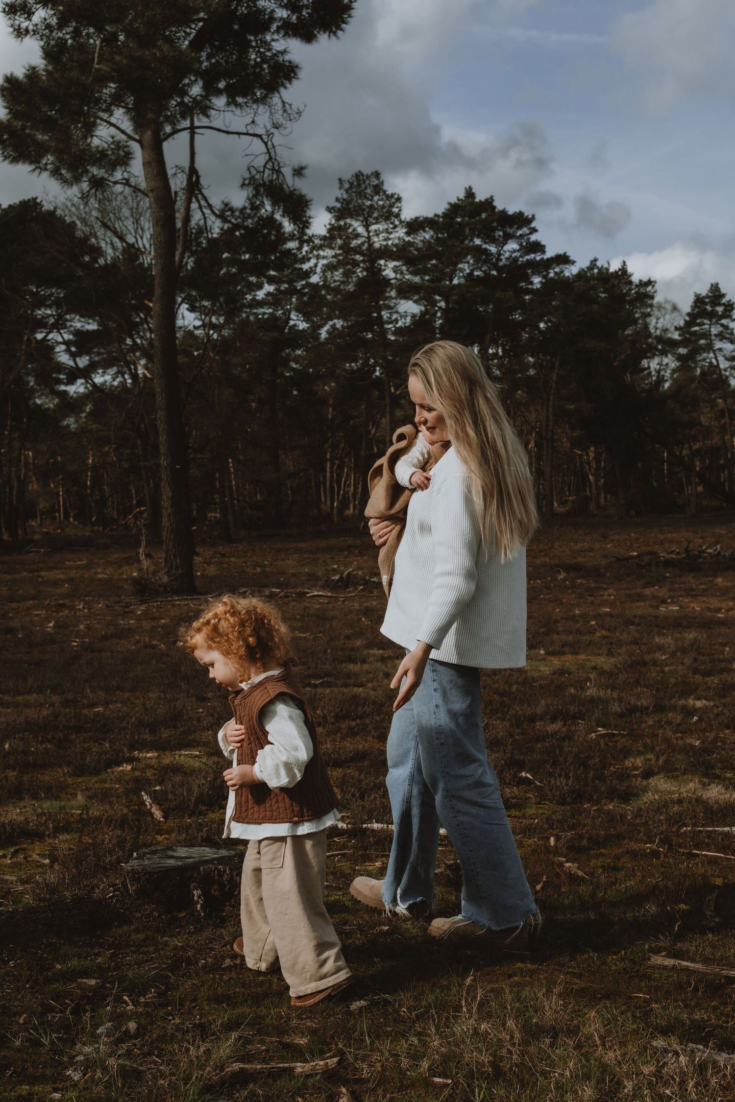 A mother carrying a baby walks alongside her curly-haired toddler in a forest.
