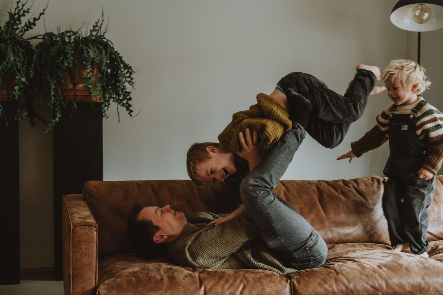 Father playfully lifting child on couch while toddler stands nearby in cozy living room.