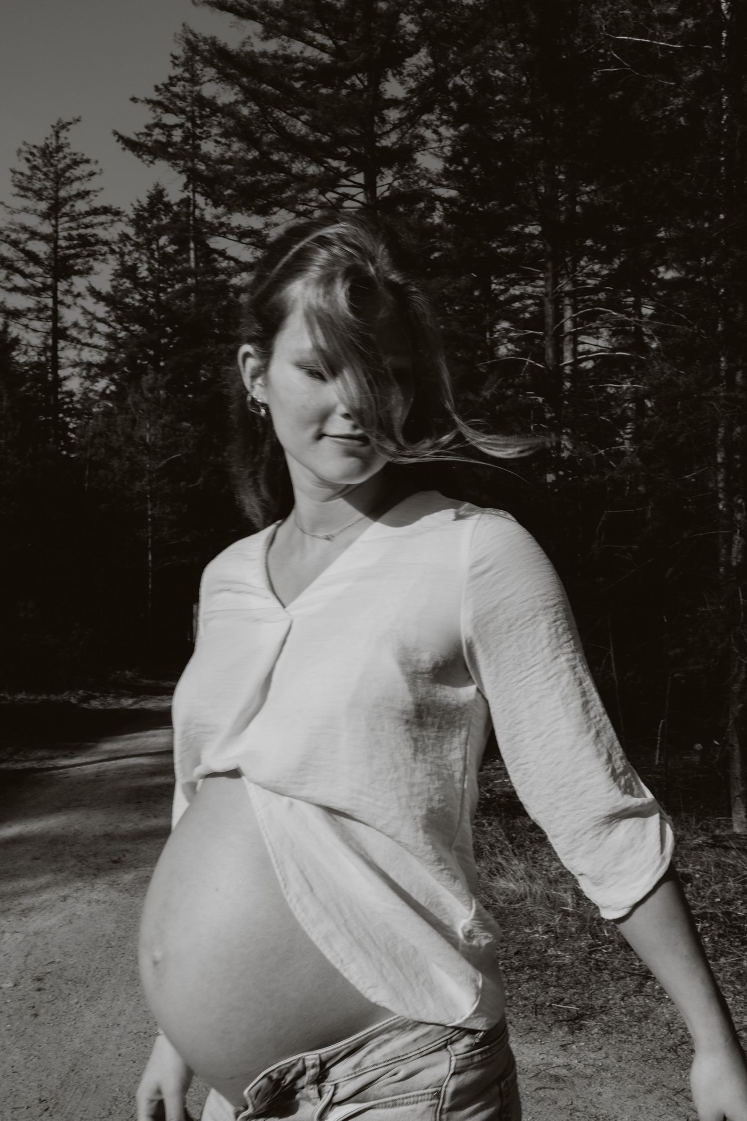 Black and white photo of pregnant woman in white blouse with wind-blown hair outdoors.