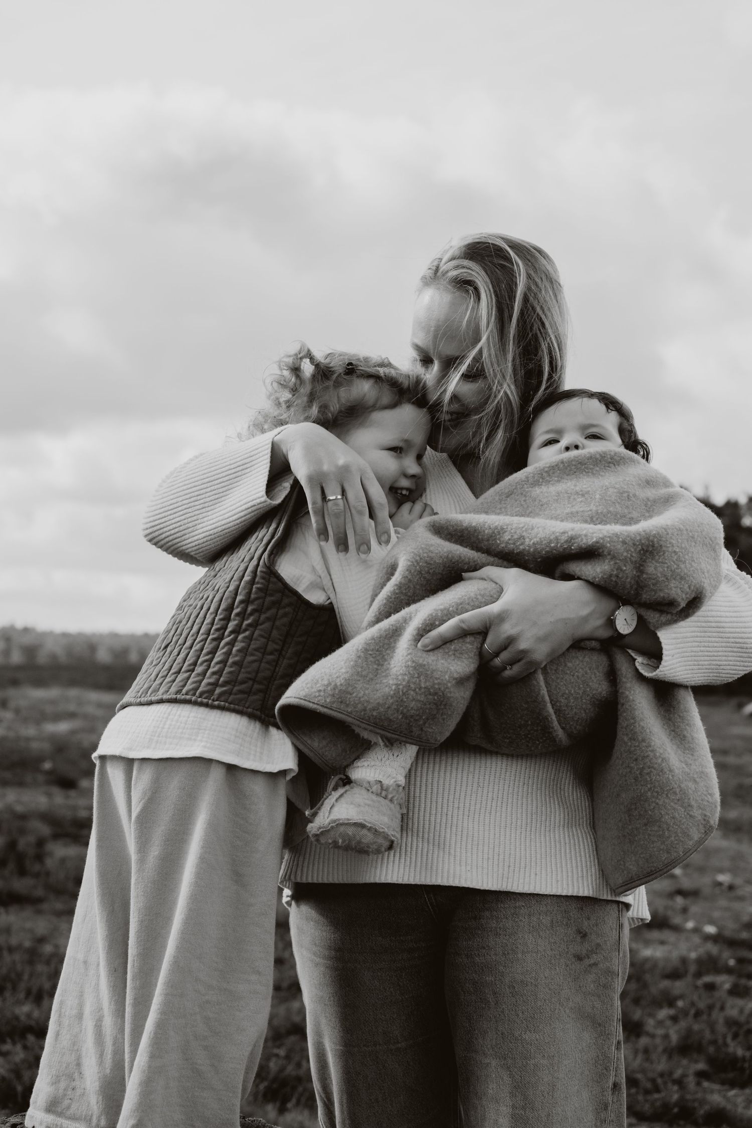 Mother embracing two young children outdoors under cloudy sky in black and white.