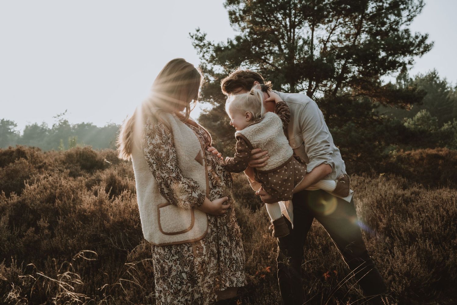 A family with a pregnant mother, father, and toddler share a tender moment in golden sunlight.