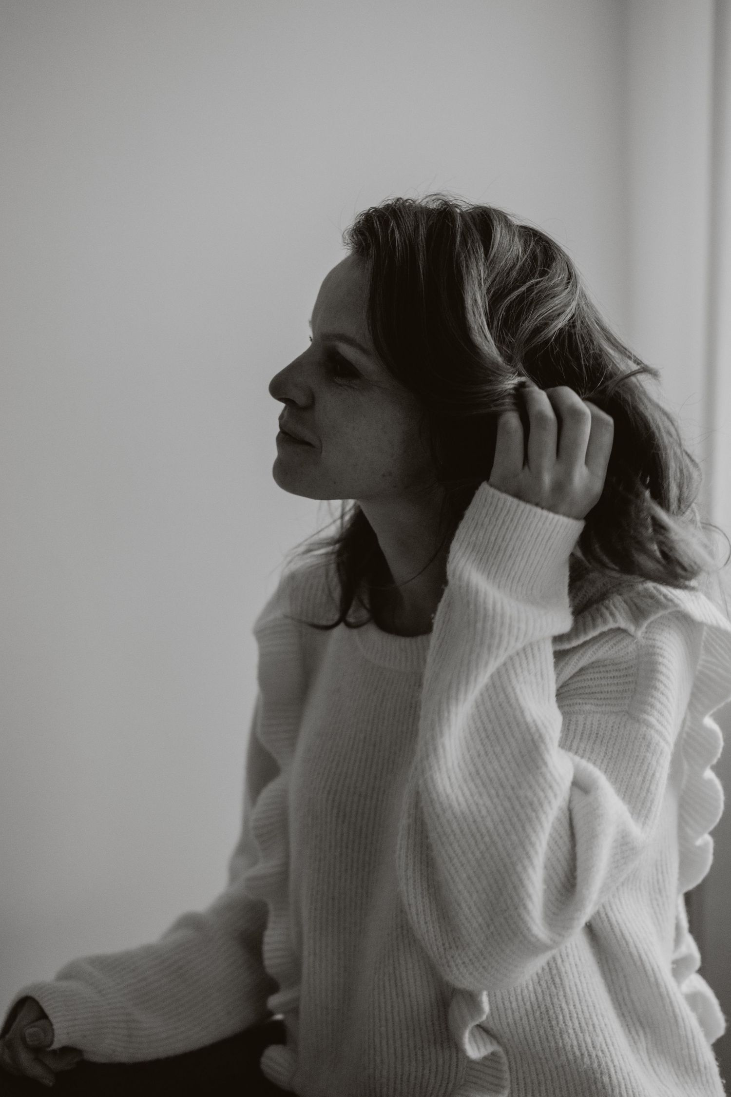 Black and white portrait of a woman in white sweater touching her hair by a window.