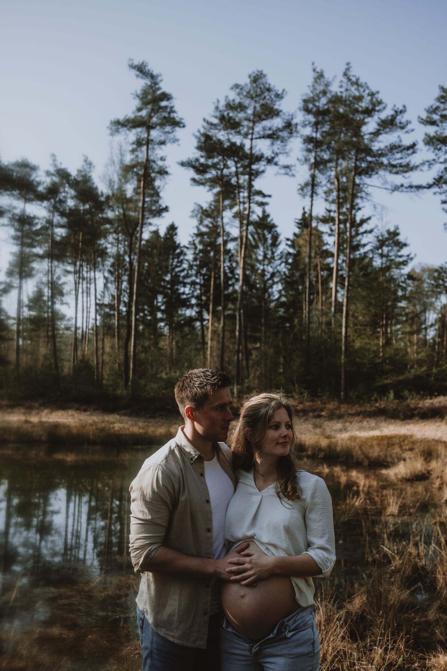 Expecting couple embracing near reflective forest pond, man holding pregnant woman's belly, tall pine trees behind.