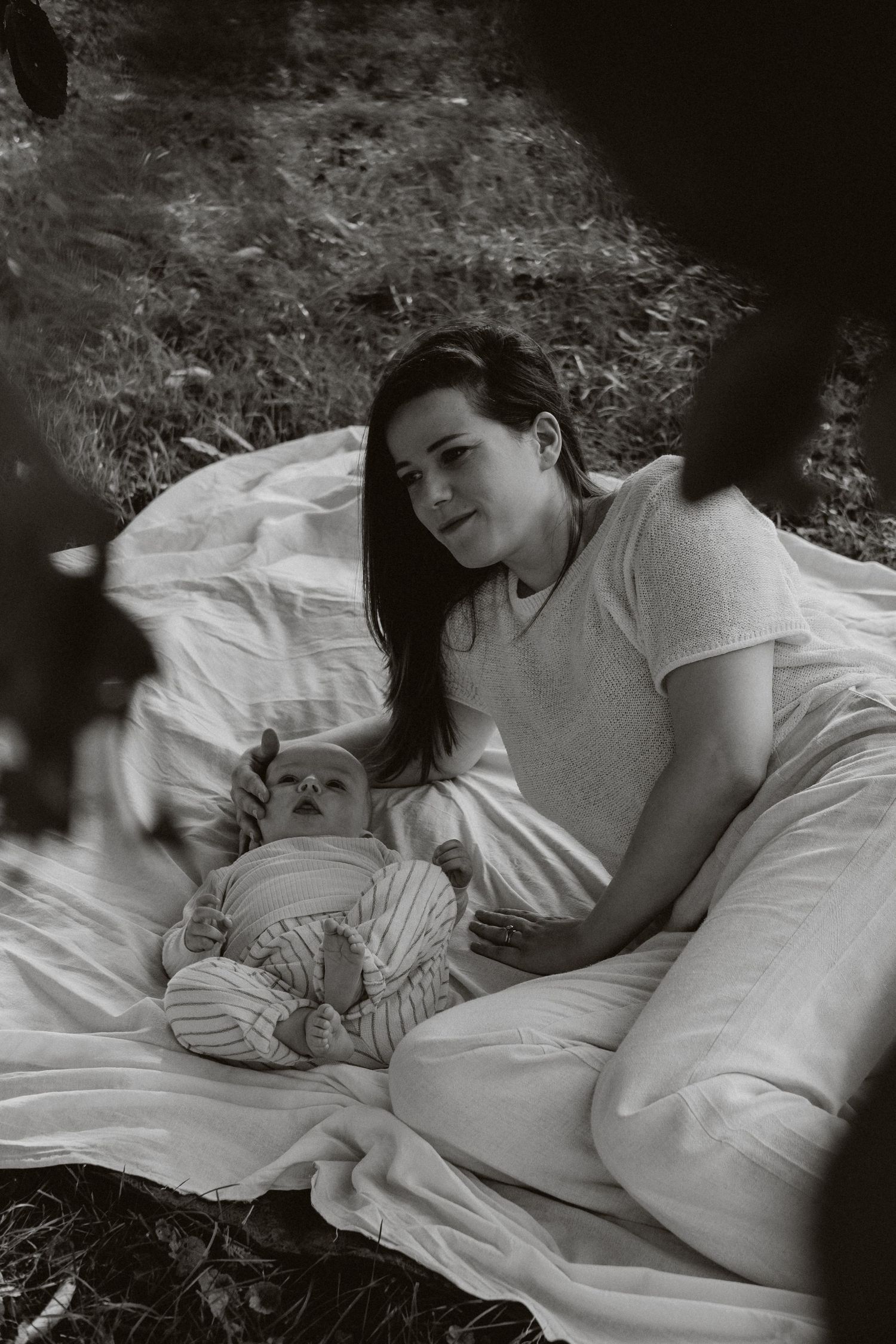 Mother relaxing on white blanket outdoors with baby in black and white portrait.