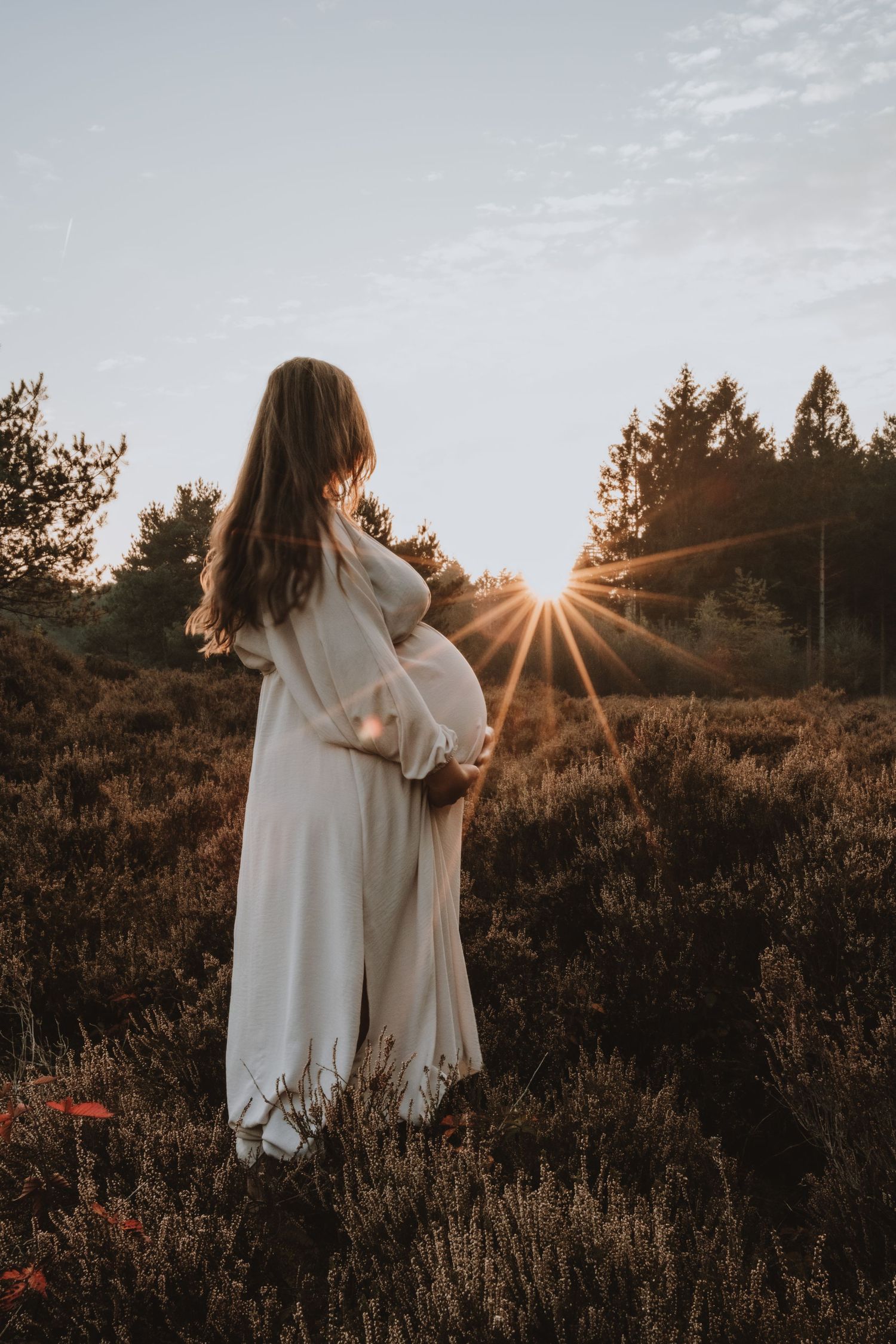 A pregnant woman in a white dress stands in a heathland field at golden sunset with sun rays.