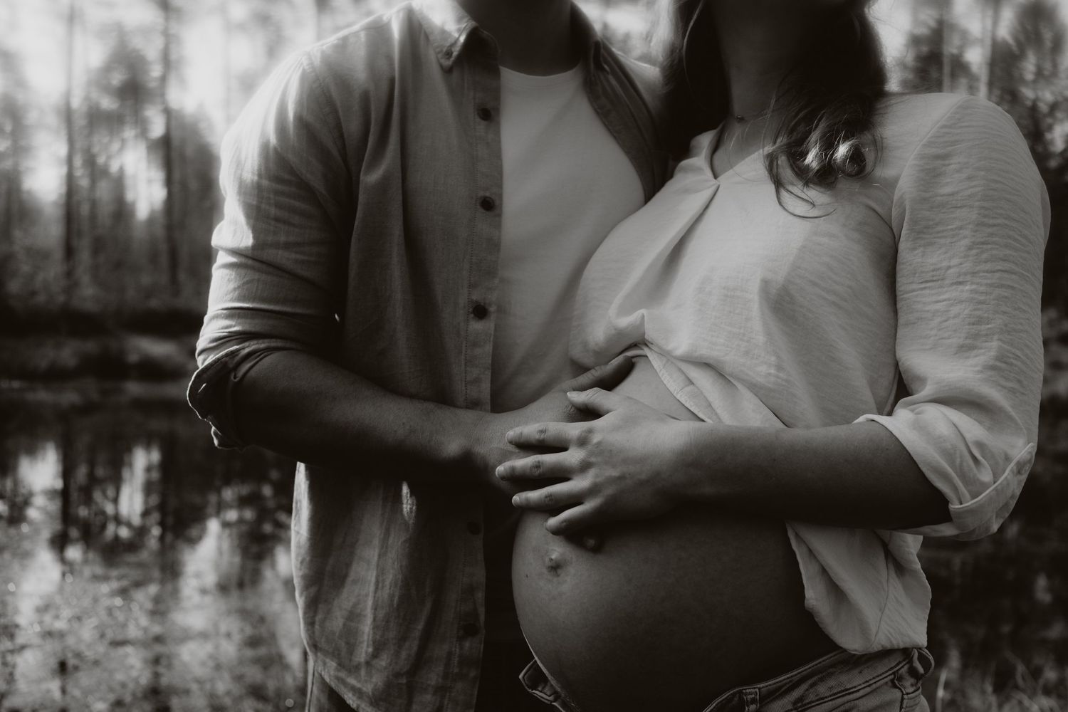 Black and white photo of couple near forest pond, man's hands cradling pregnant woman's bare belly.