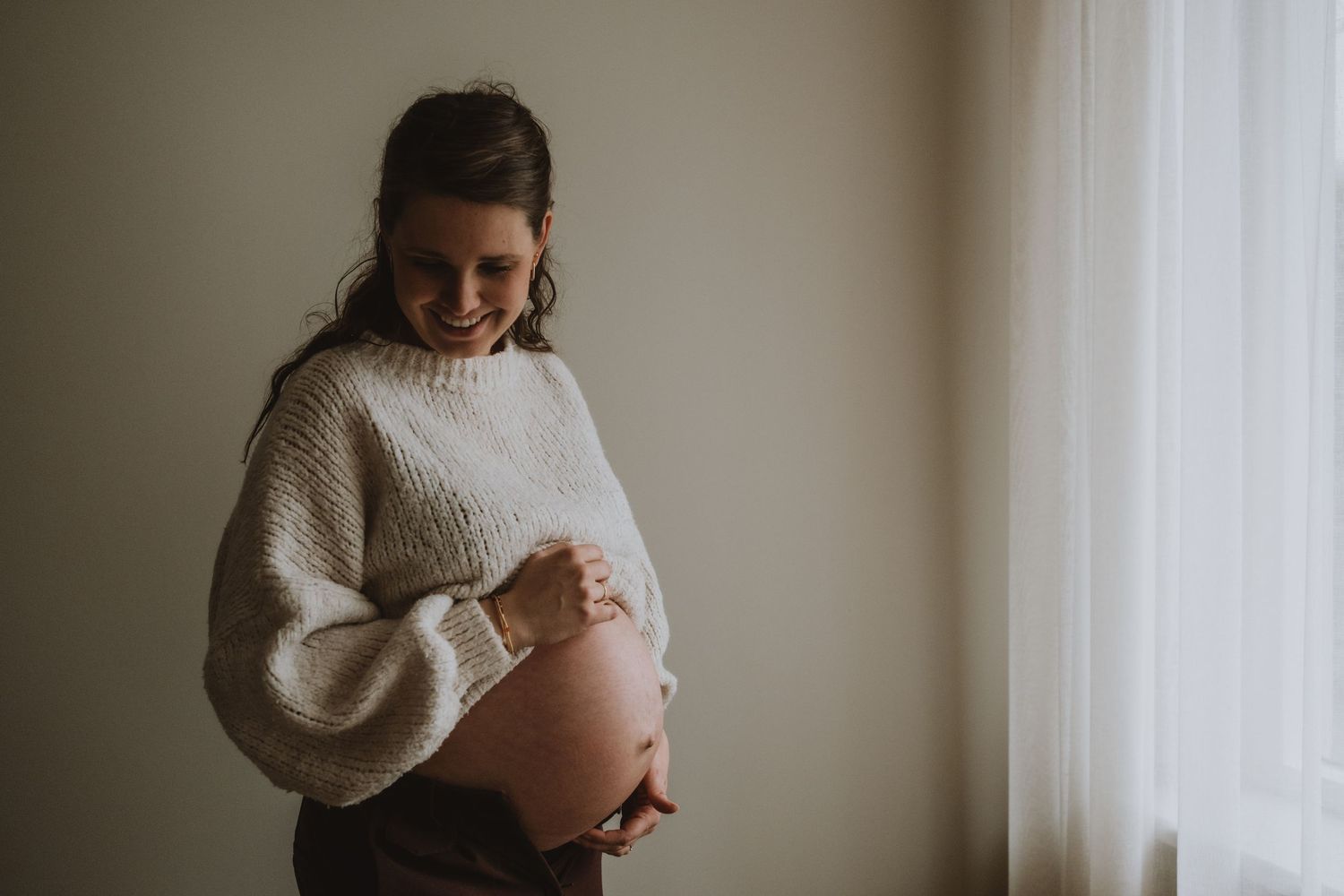 Smiling pregnant woman in cream knit sweater holding baby bump near bright window.