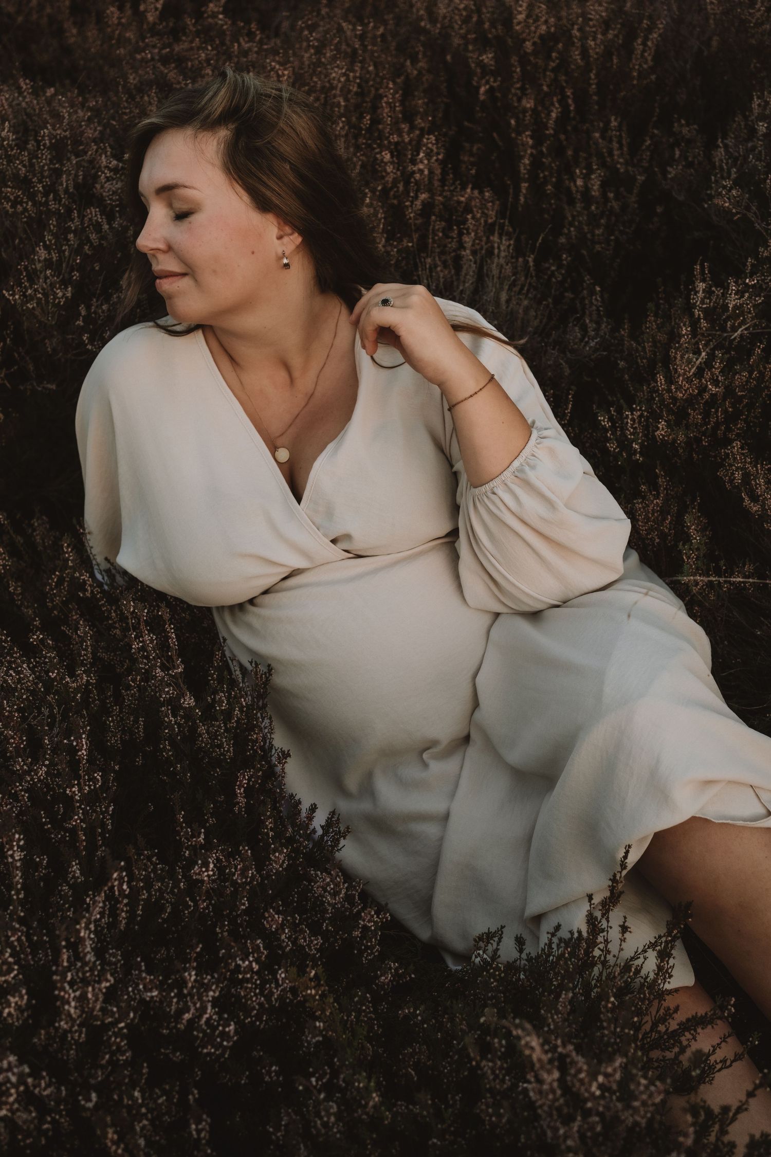 A pregnant woman in a cream dress reclines peacefully among dark heather plants outdoors.
