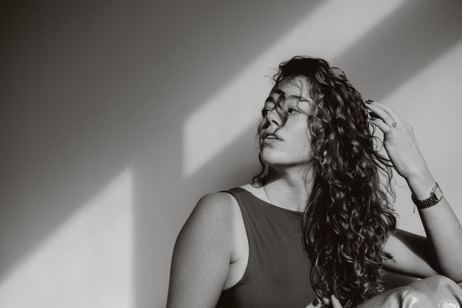 Black and white portrait of a young woman with curly hair touching her locks, eyes closed in soft natural light.
