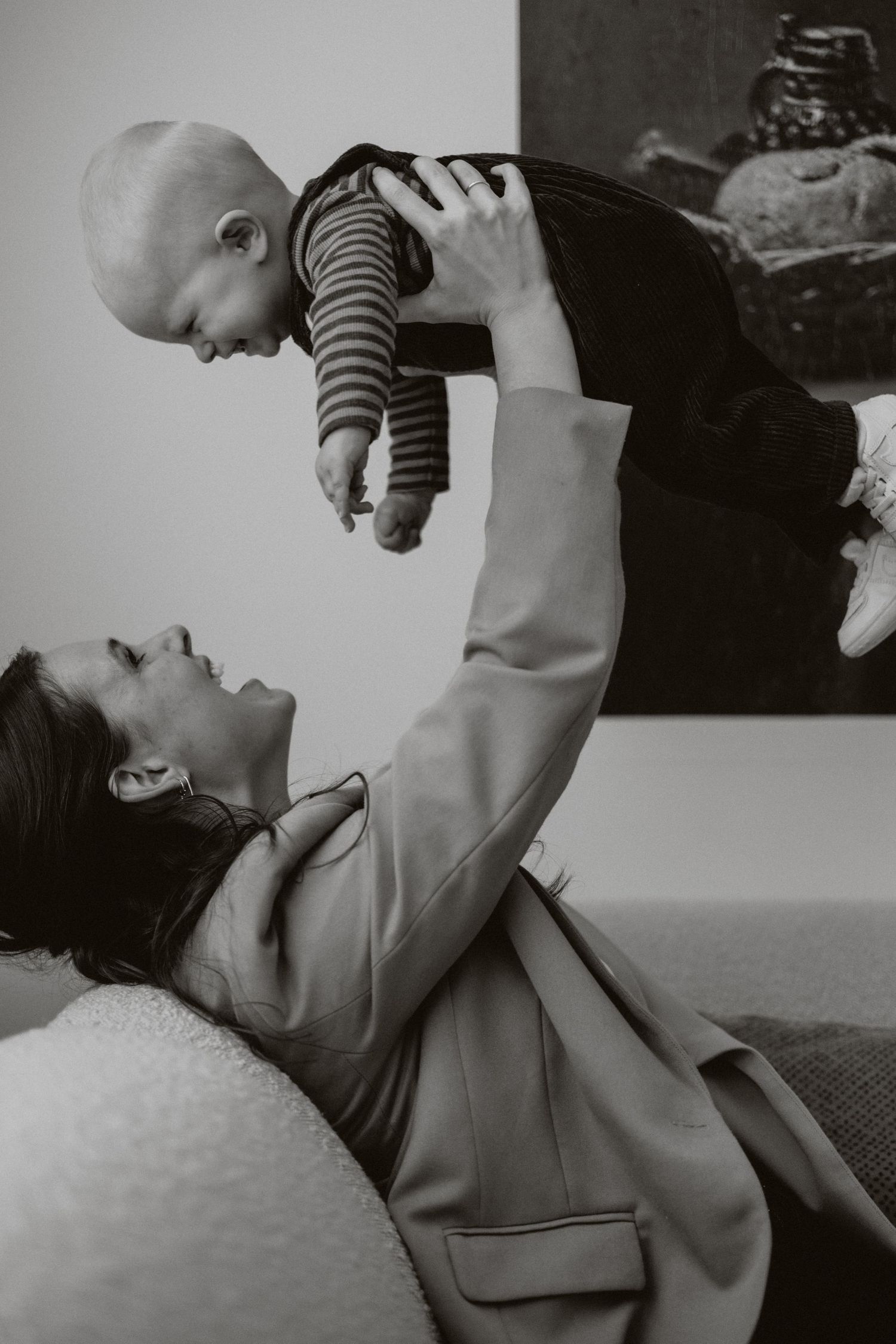 Black and white photo of a mother lying on a sofa lifting her baby up joyfully.