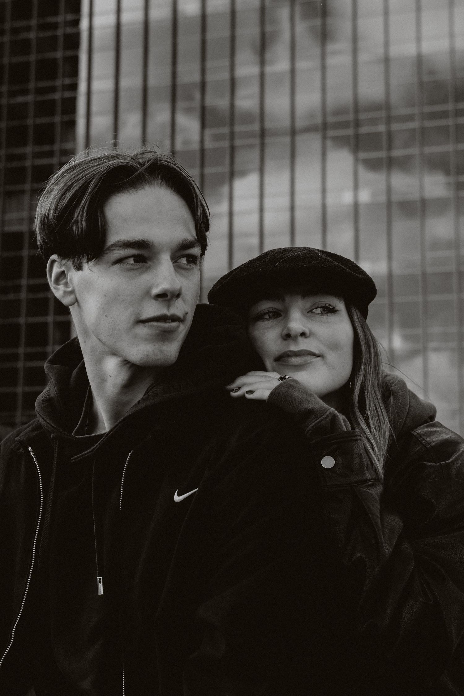 Black and white portrait of young couple posing together against metal grid fence outdoors.