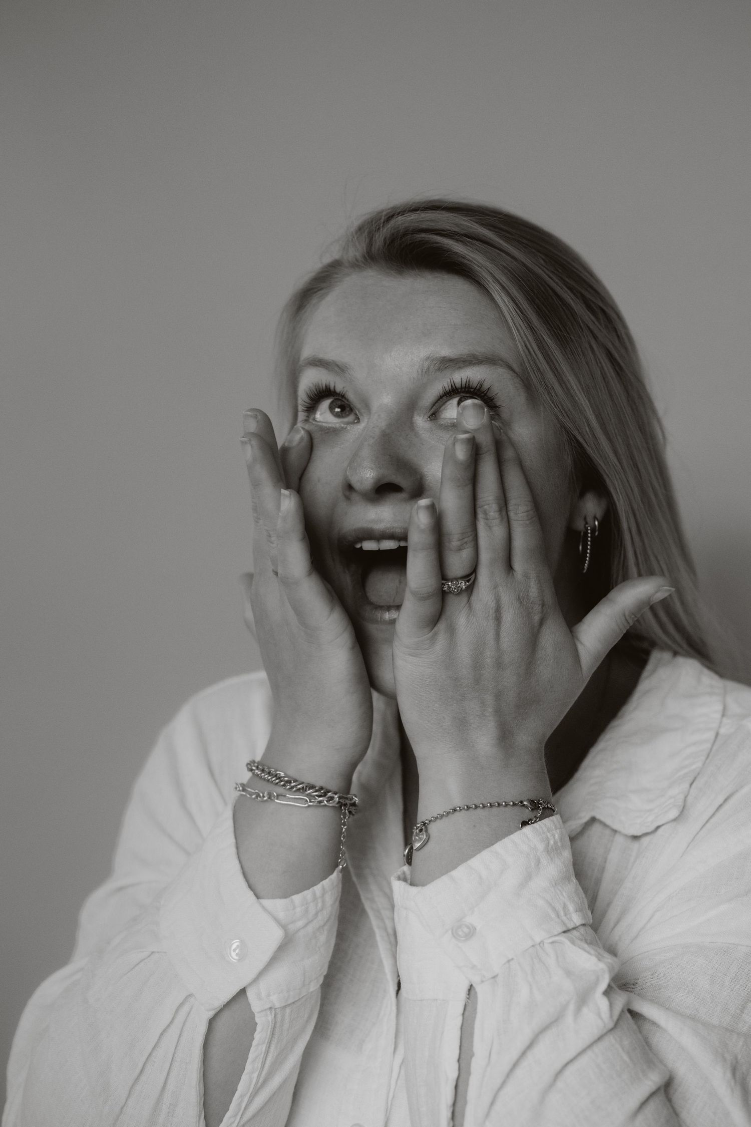 Surprised blonde woman holds hands to face in expressive black and white portrait.