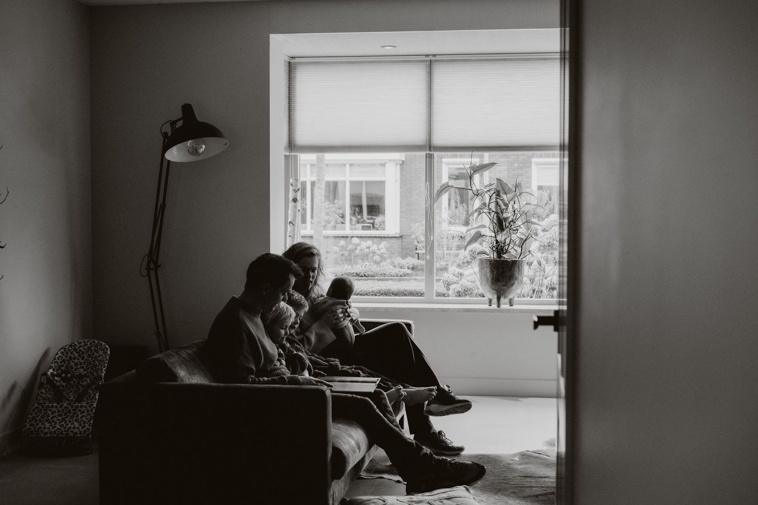 Black and white photo of a family sitting together on a sofa near a bright window in a cozy living room.