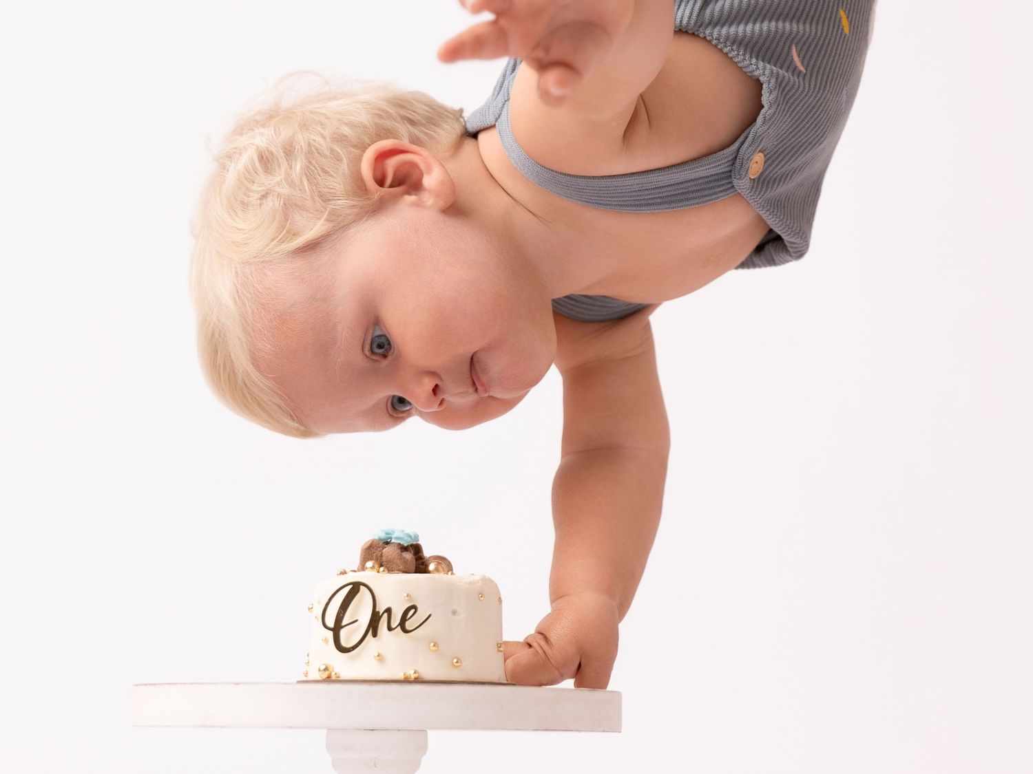 Blonde toddler leaning over a small white cake decorated with gold One text during a first birthday smash cake photoshoot.
