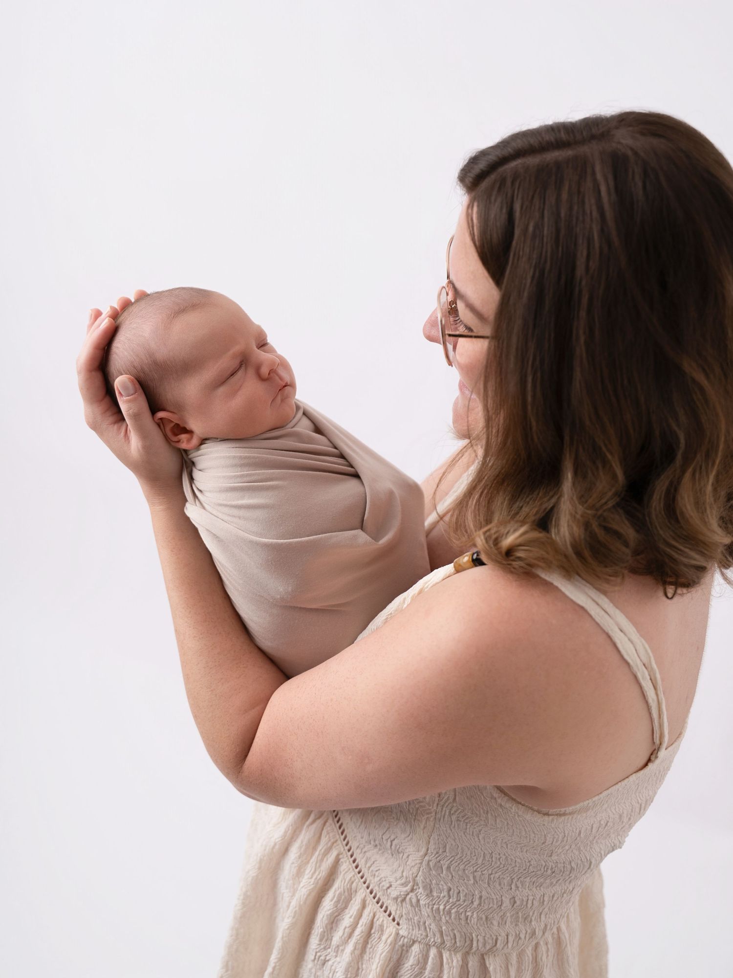 A mother in a beige dress holds her swaddled newborn baby, gazing lovingly at the infant against a white background.