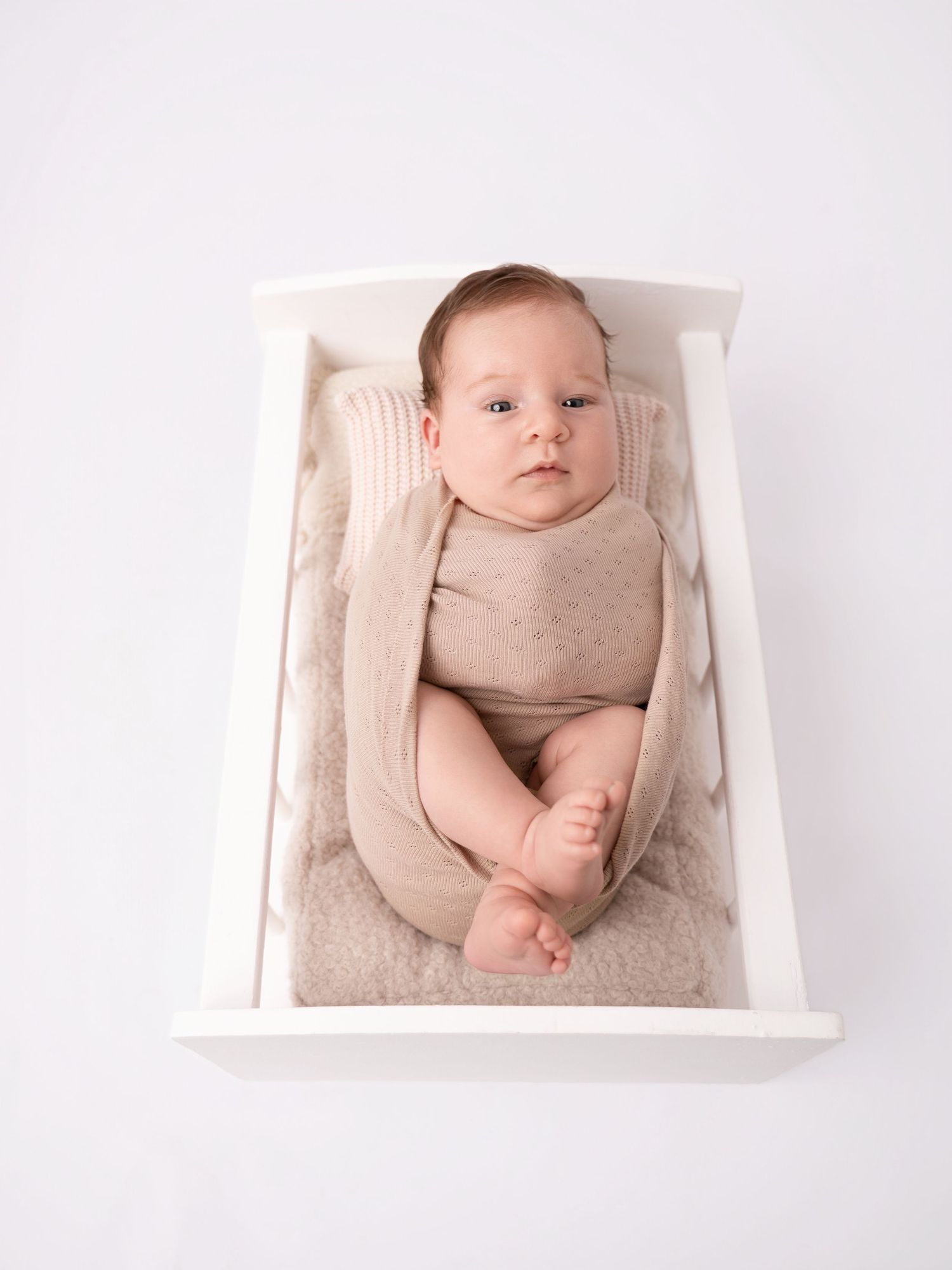 Newborn baby in beige outfit lying in a white wooden doll bed on a white background.