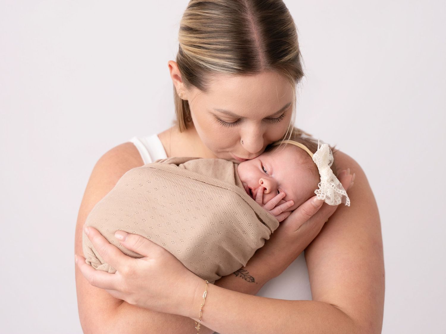 A mother tenderly holds her swaddled newborn baby wrapped in a beige blanket against a white background.
