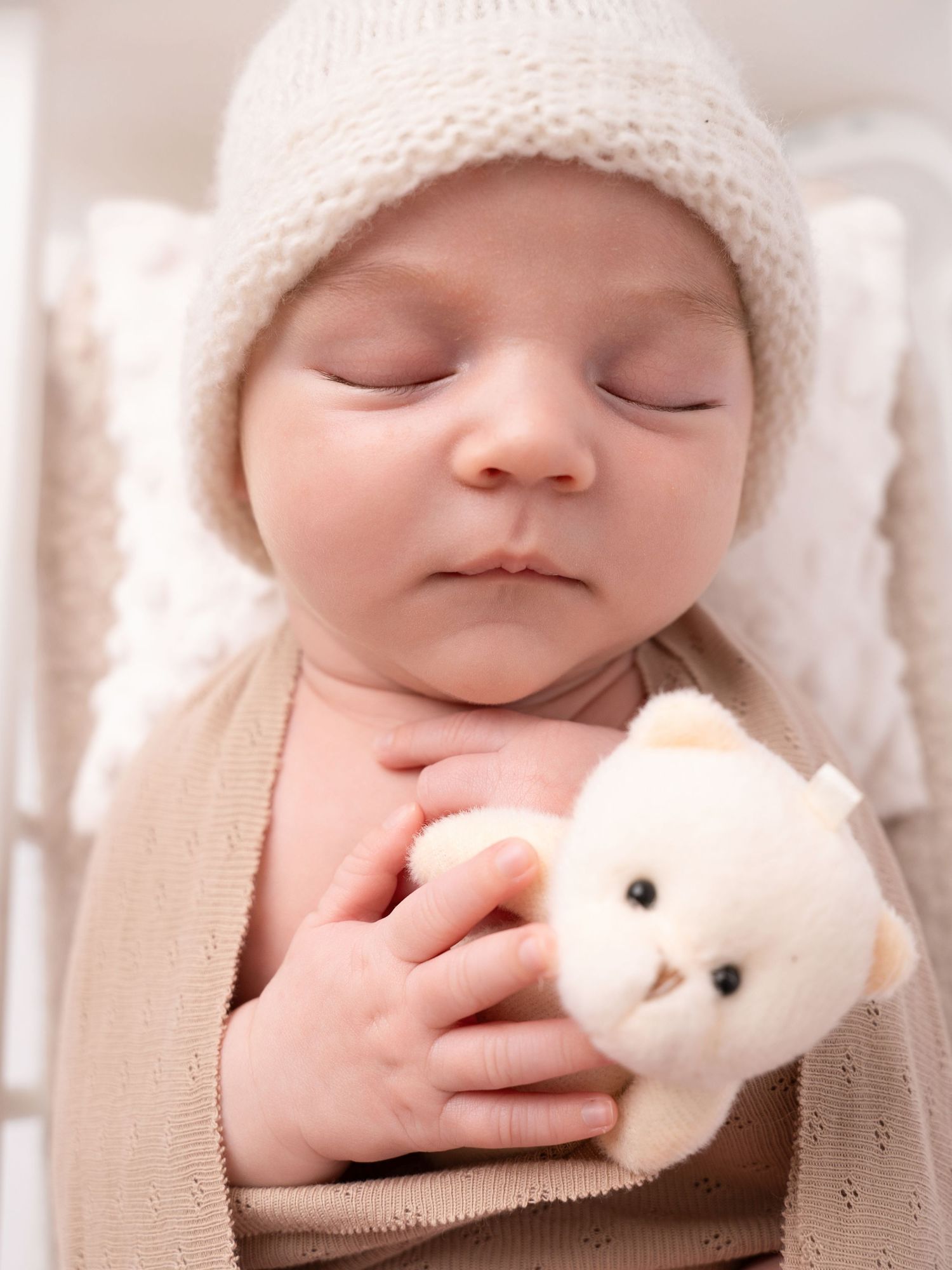 Sleeping newborn baby in a knit hat holds a tiny white teddy bear, wrapped in a soft beige blanket.
