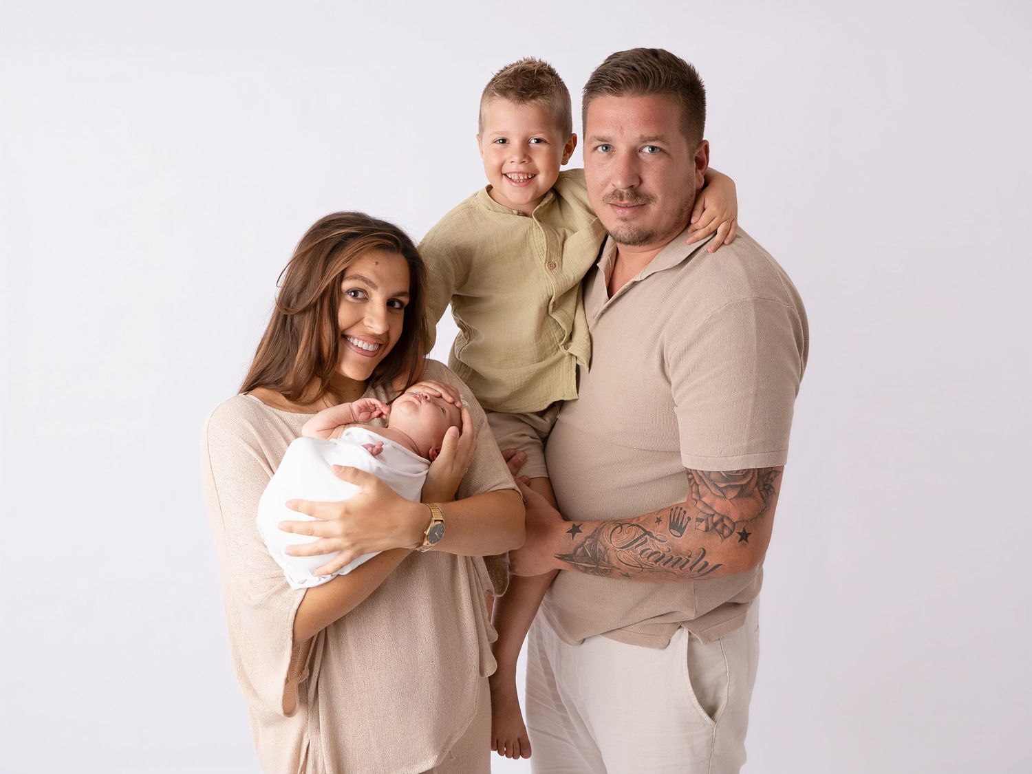 A happy family of four poses for a professional portrait, with parents holding a newborn and toddler against a white background.