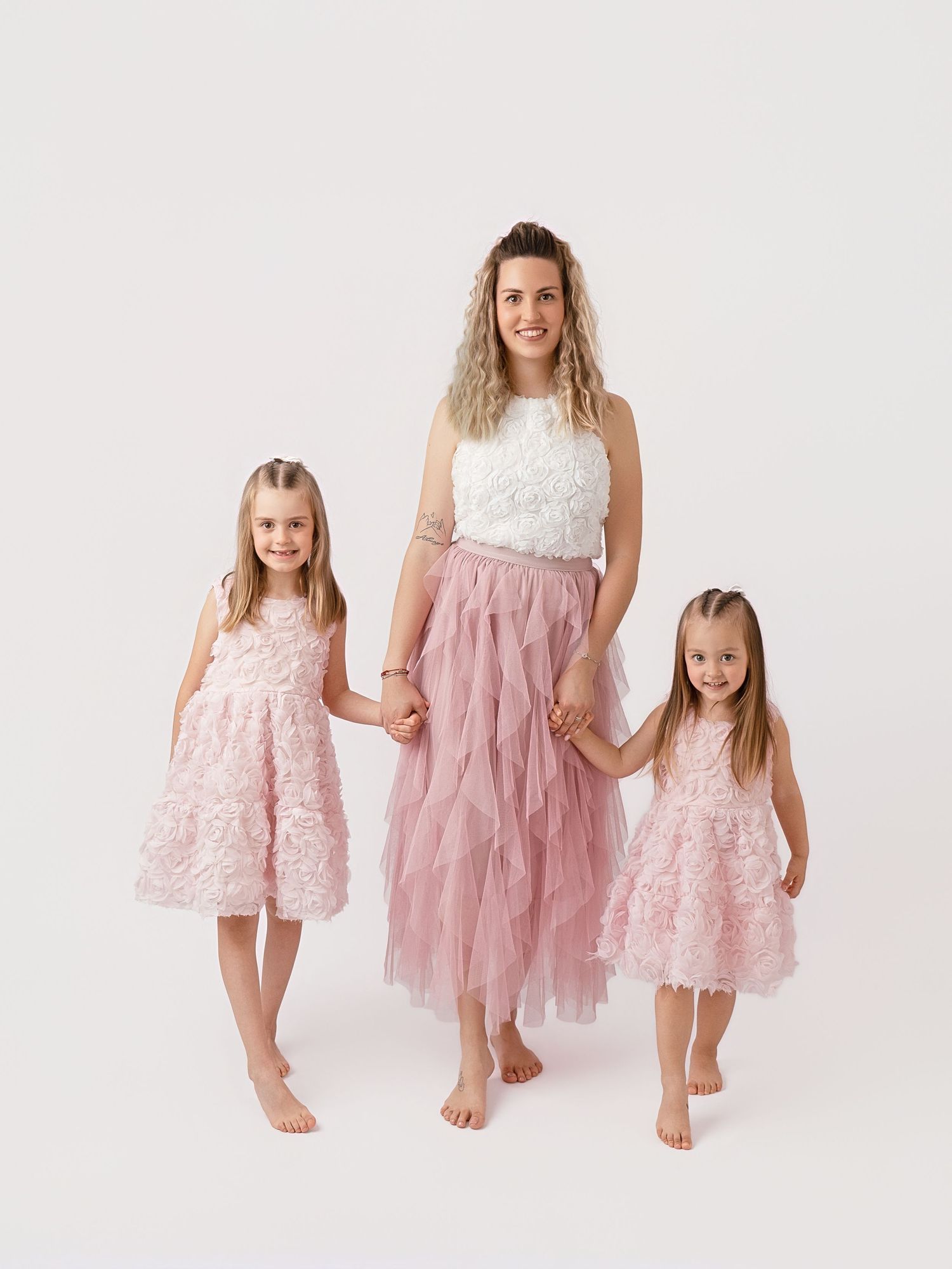 A mother and two daughters in matching pink floral dresses pose together holding hands on a white background.