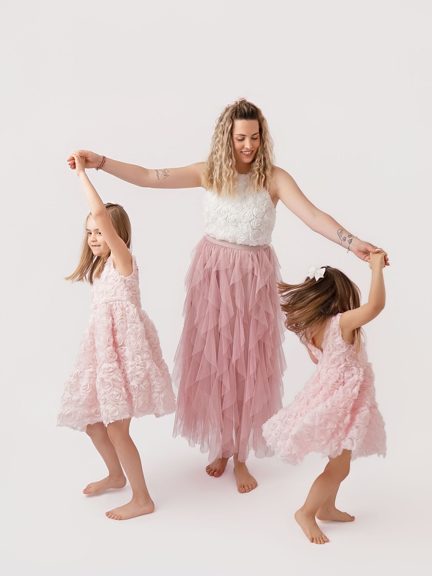 Woman and two girls in matching pink tulle dresses dancing together on white background.