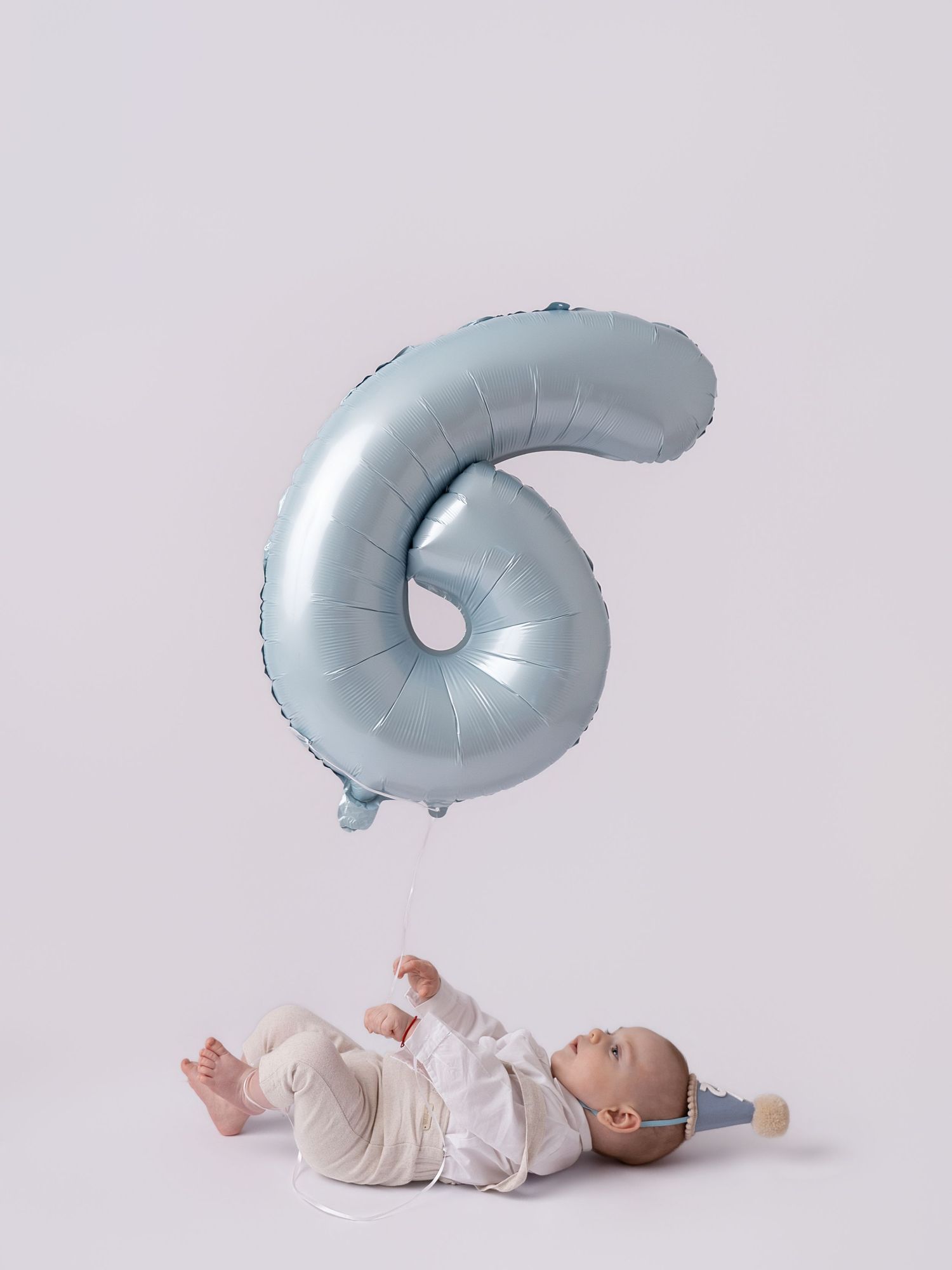 Baby lying on back holding a light blue number 6 foil balloon, celebrating six months old milestone.