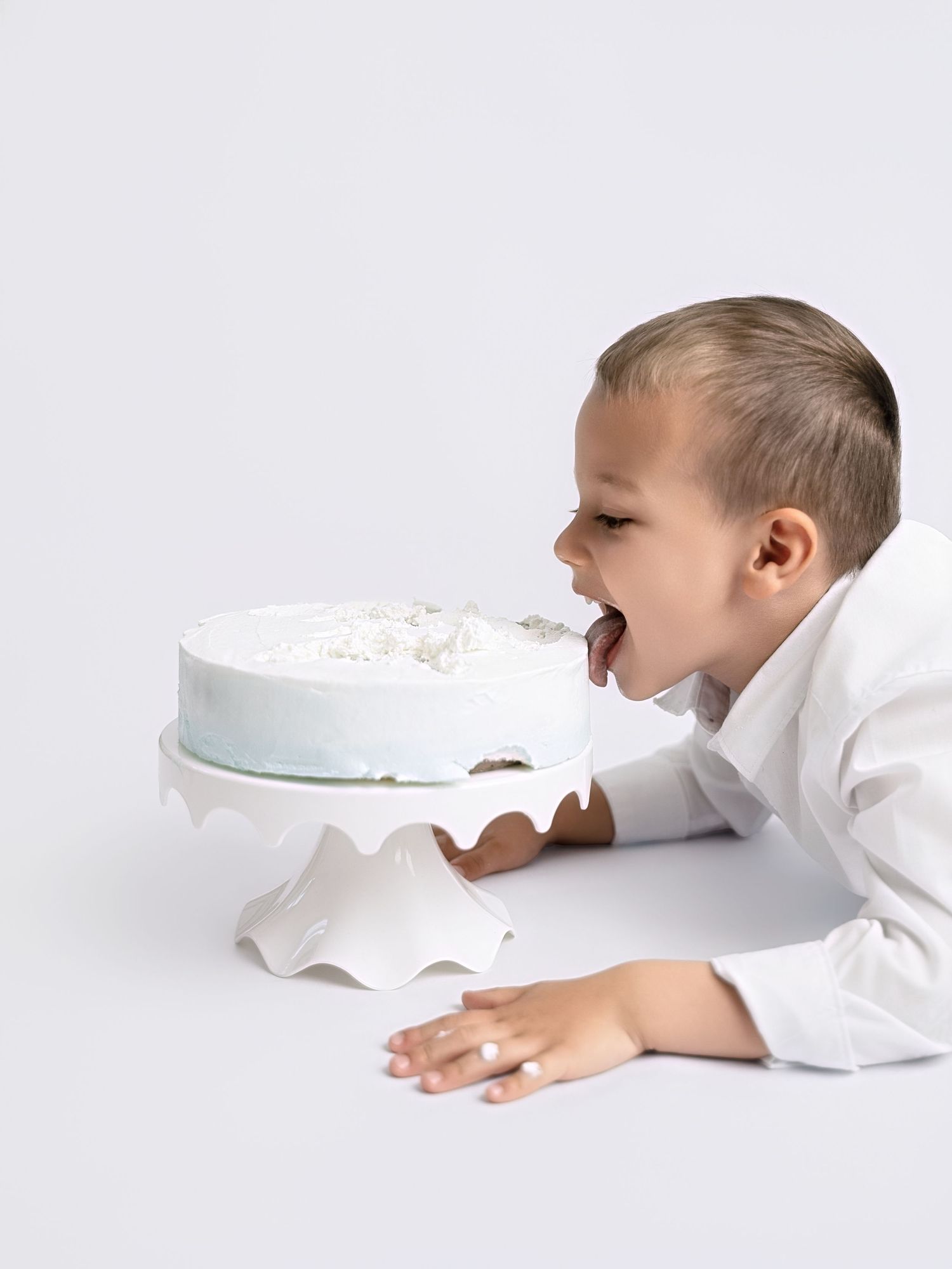 Young boy in white shirt eagerly biting into a white frosted cake on a decorative cake stand.