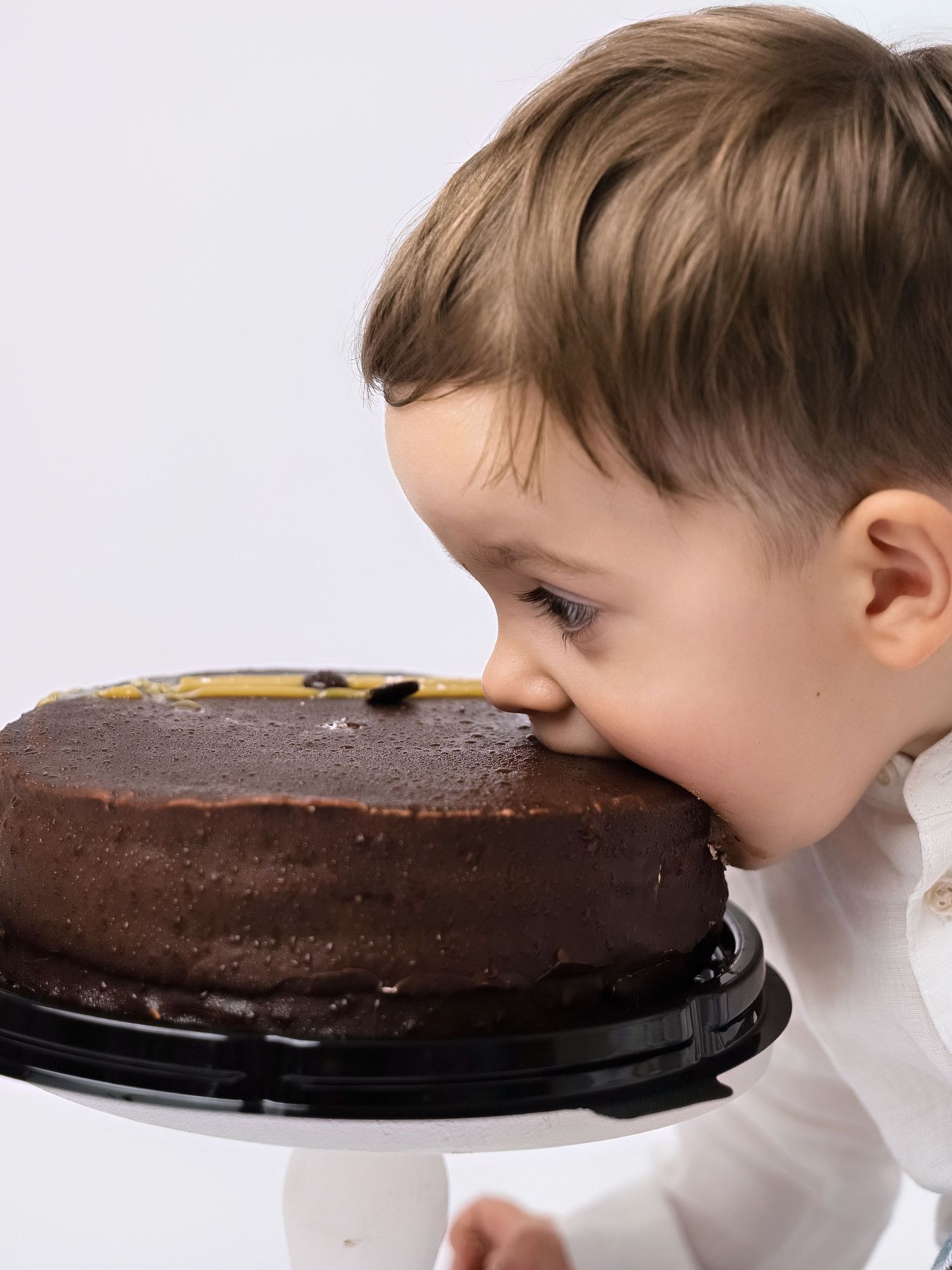 Toddler boy in white shirt taking a big bite out of a chocolate frosted birthday cake on a white stand.