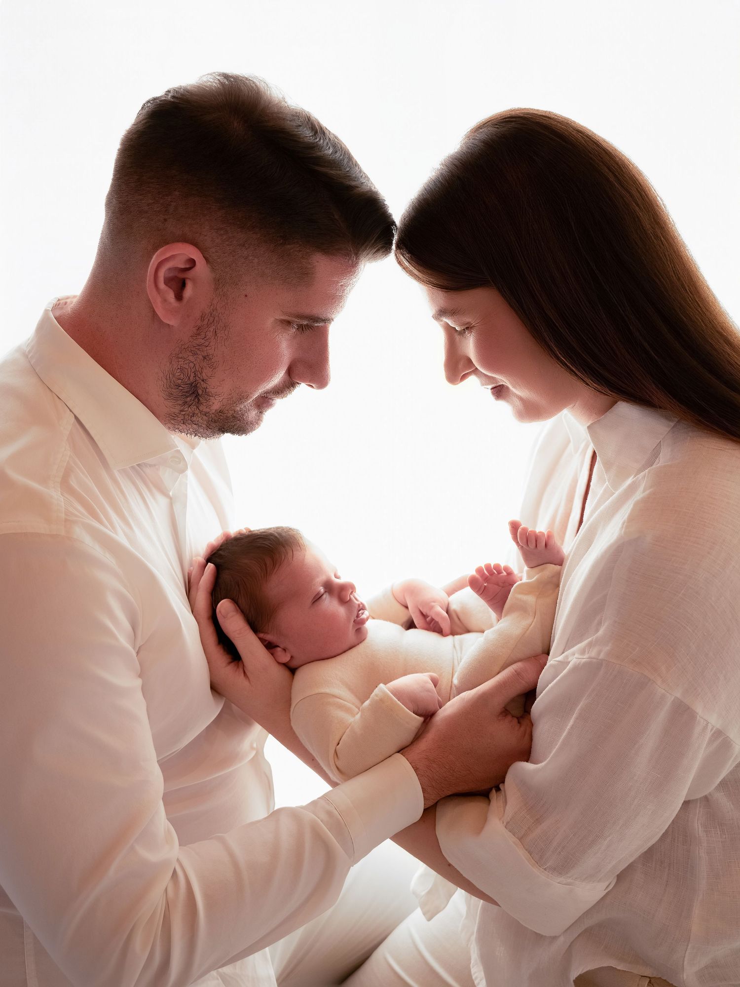 A couple in white clothing tenderly cradles their newborn baby together in a soft, bright studio portrait.