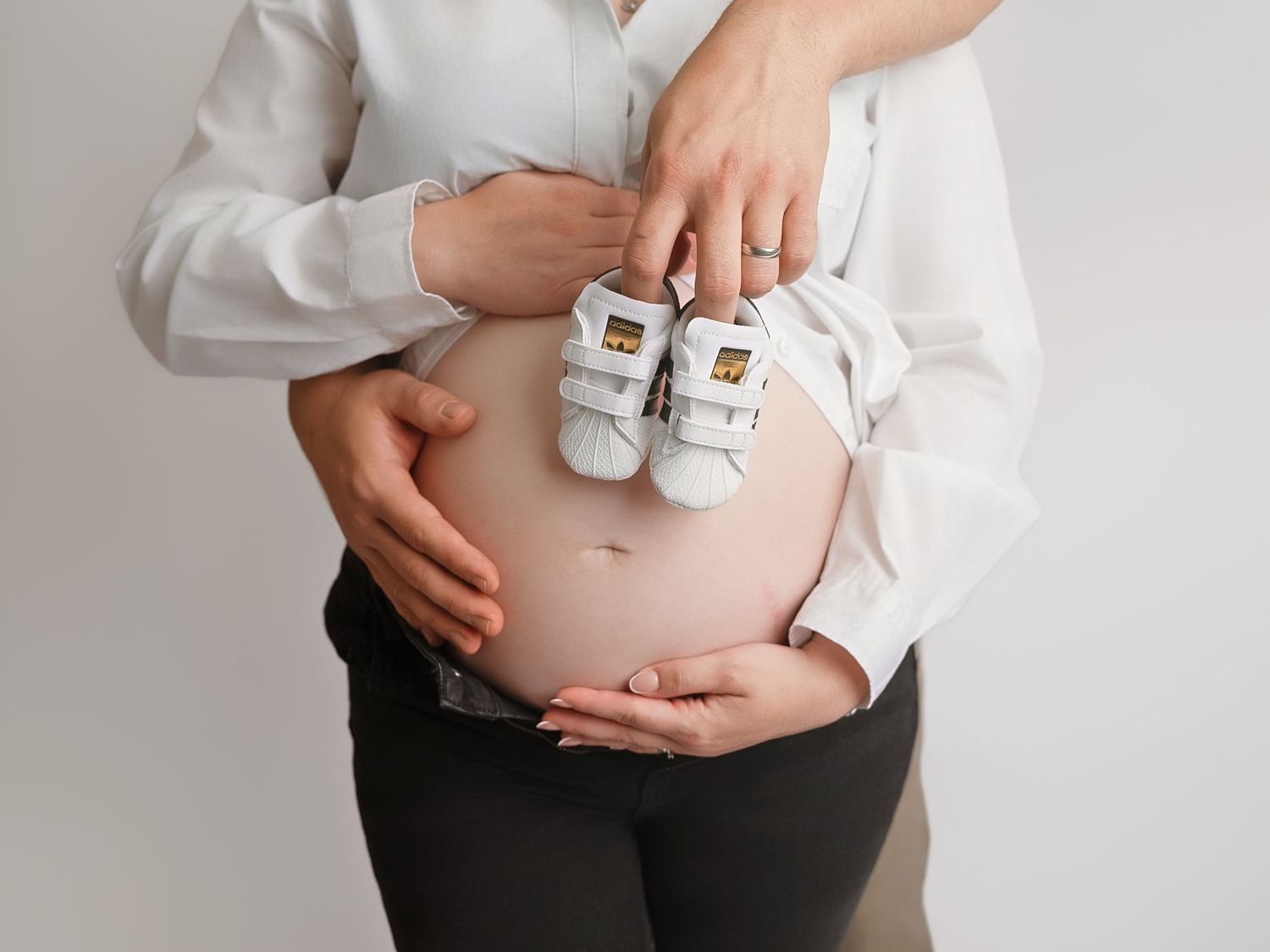 Pregnant woman holding tiny white baby sneakers over her bare belly, with a partner's hands gently cradling her bump.
