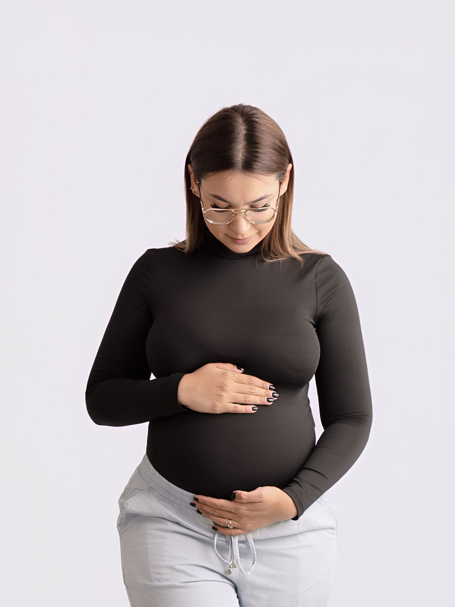 Pregnant woman in black long-sleeve top and gray pants cradling her baby bump while looking down.