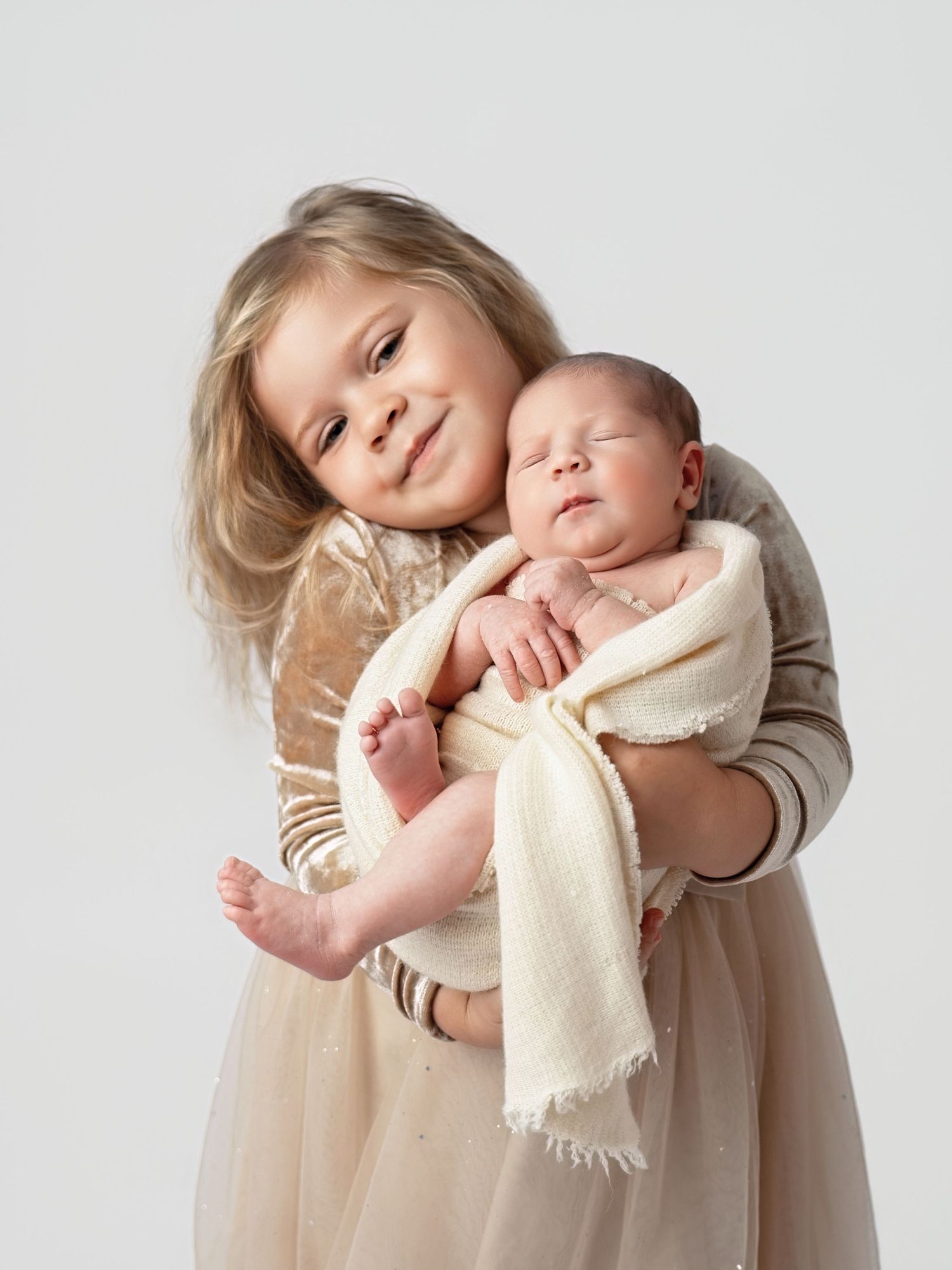 Young girl in cream dress proudly holds sleeping newborn baby wrapped in white blanket for a studio portrait.