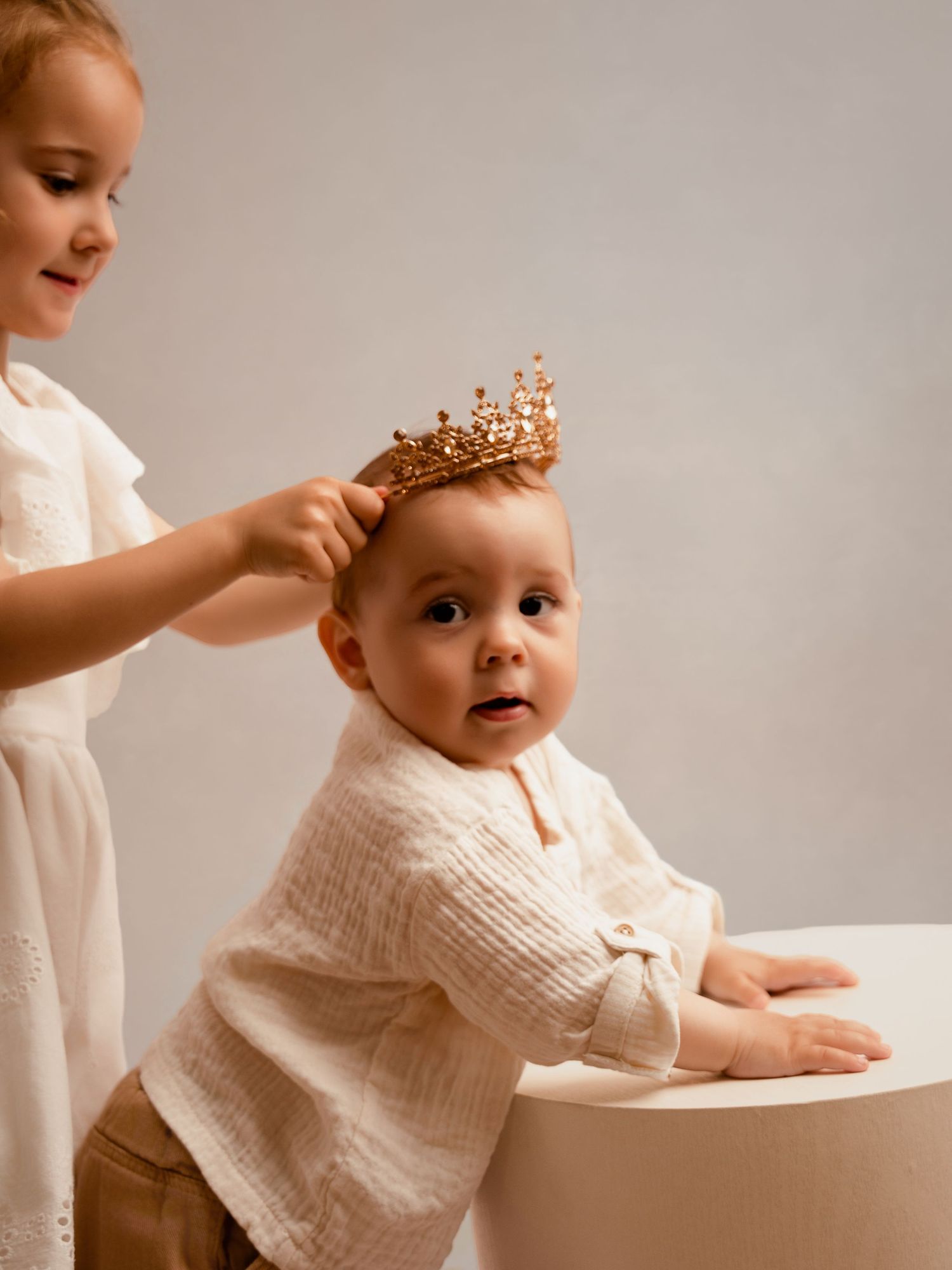 Older toddler placing a gold crown on a baby's head during a studio photo session.
