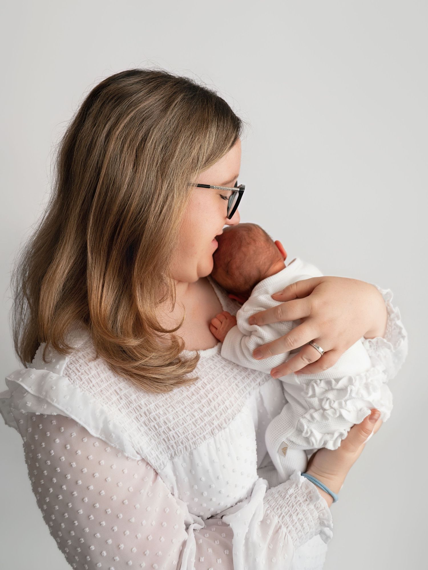 A young mother with glasses tenderly kisses her newborn baby wrapped in white, against a soft gray background.
