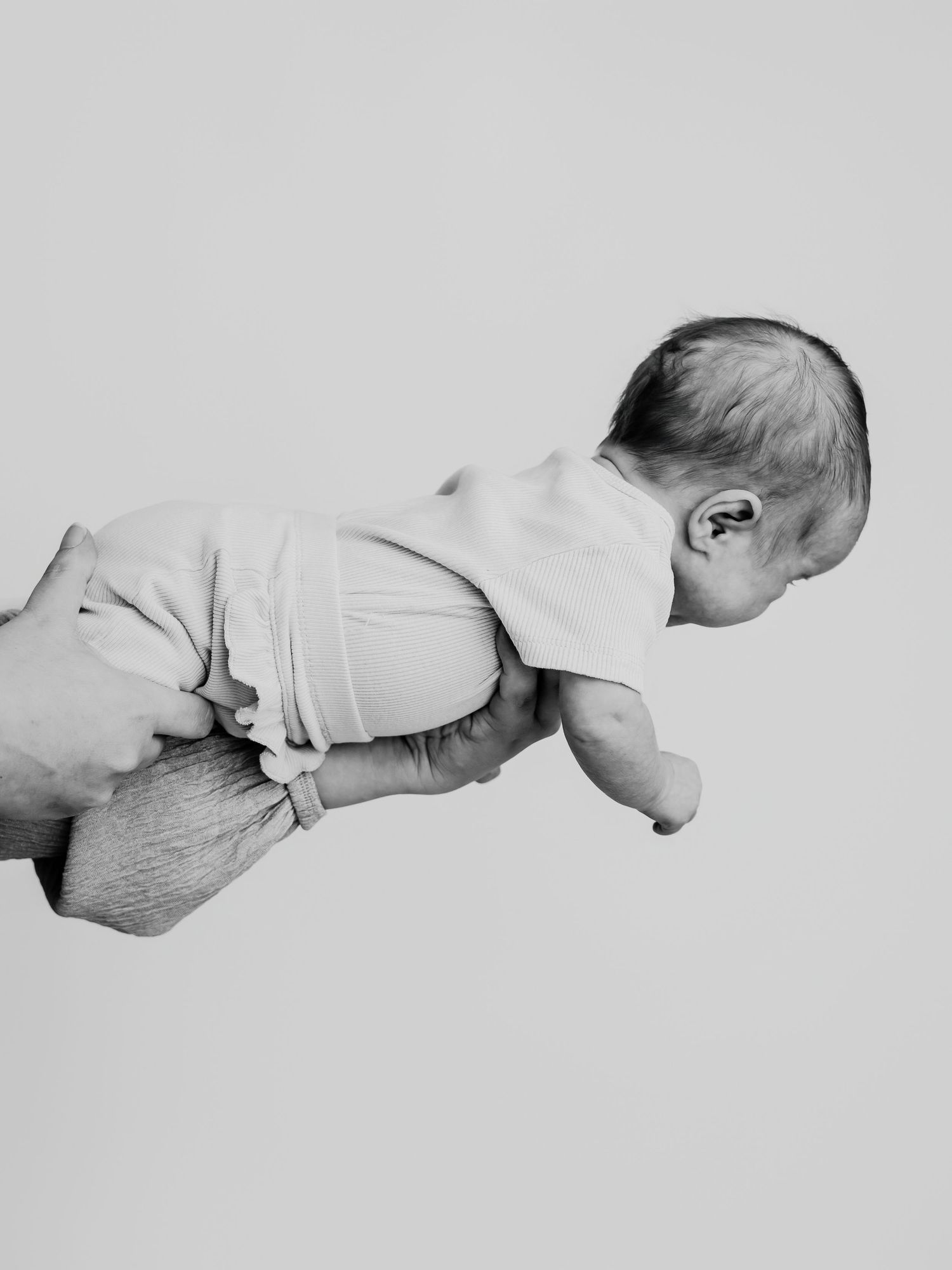 Black and white photo of newborn baby being held up by adult hands against plain background.