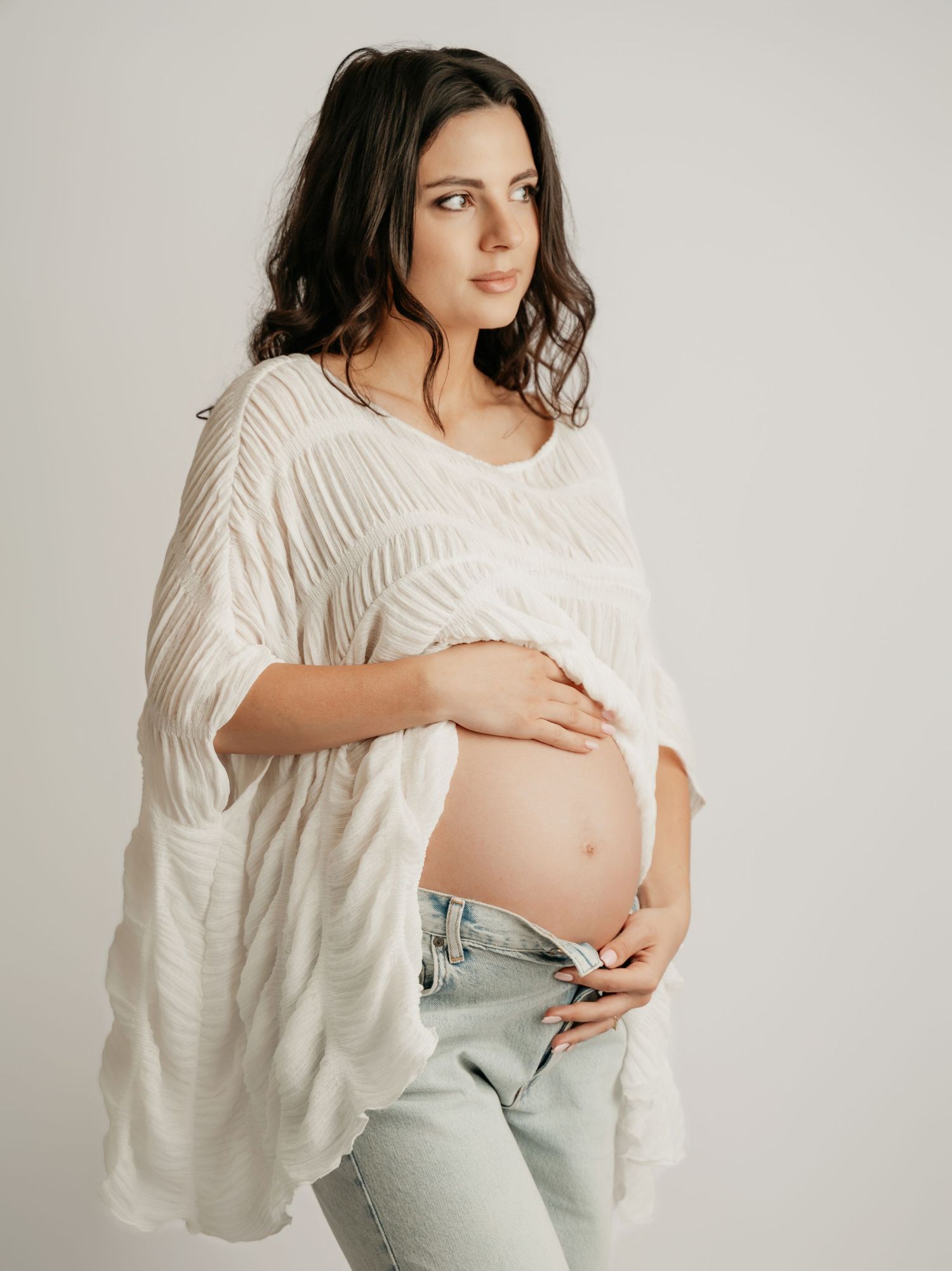 Smiling pregnant woman in white textured top and jeans holds bare baby bump in studio.
