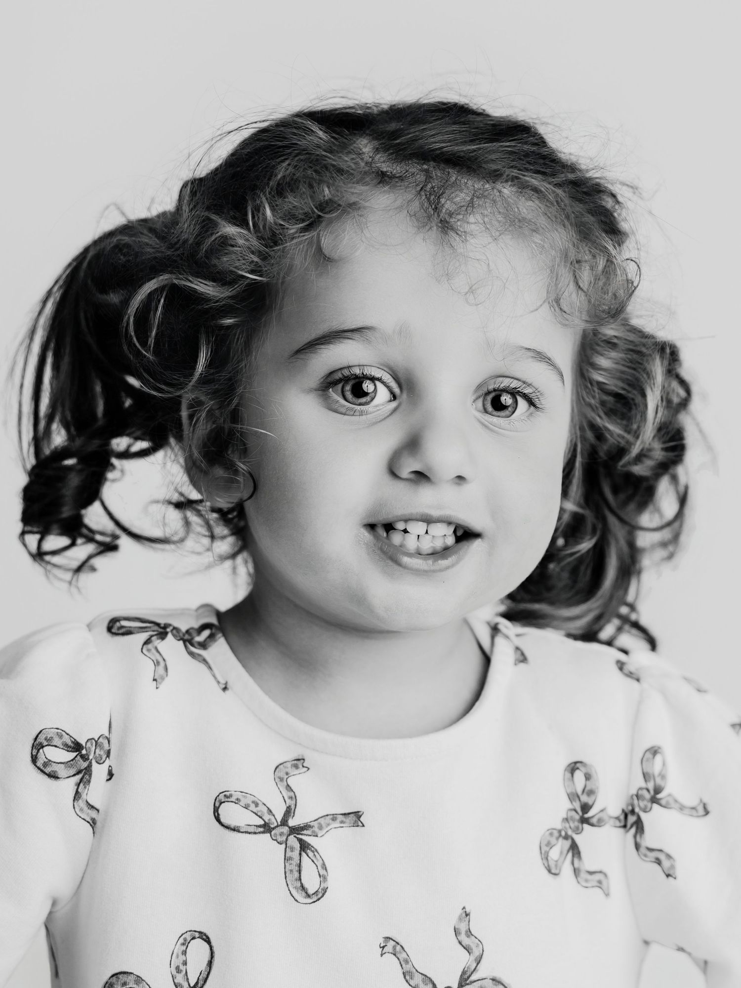 Black and white portrait of a cute curly-haired toddler girl in a bow-print white dress.