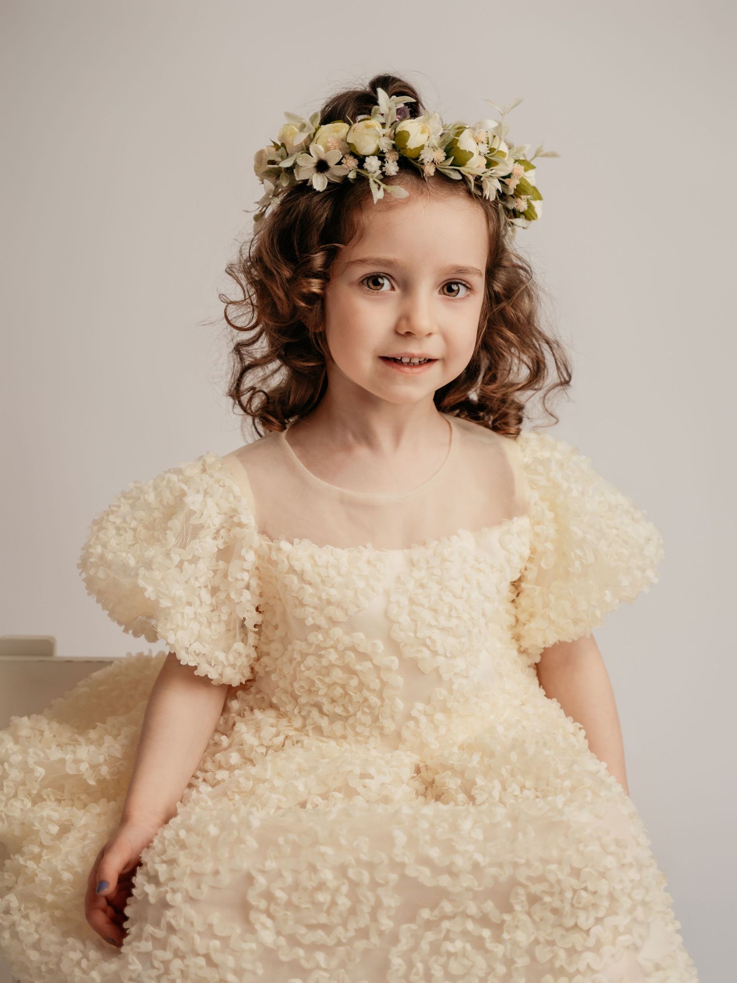 A young girl in a cream floral textured puff-sleeve dress wearing a delicate white flower crown.