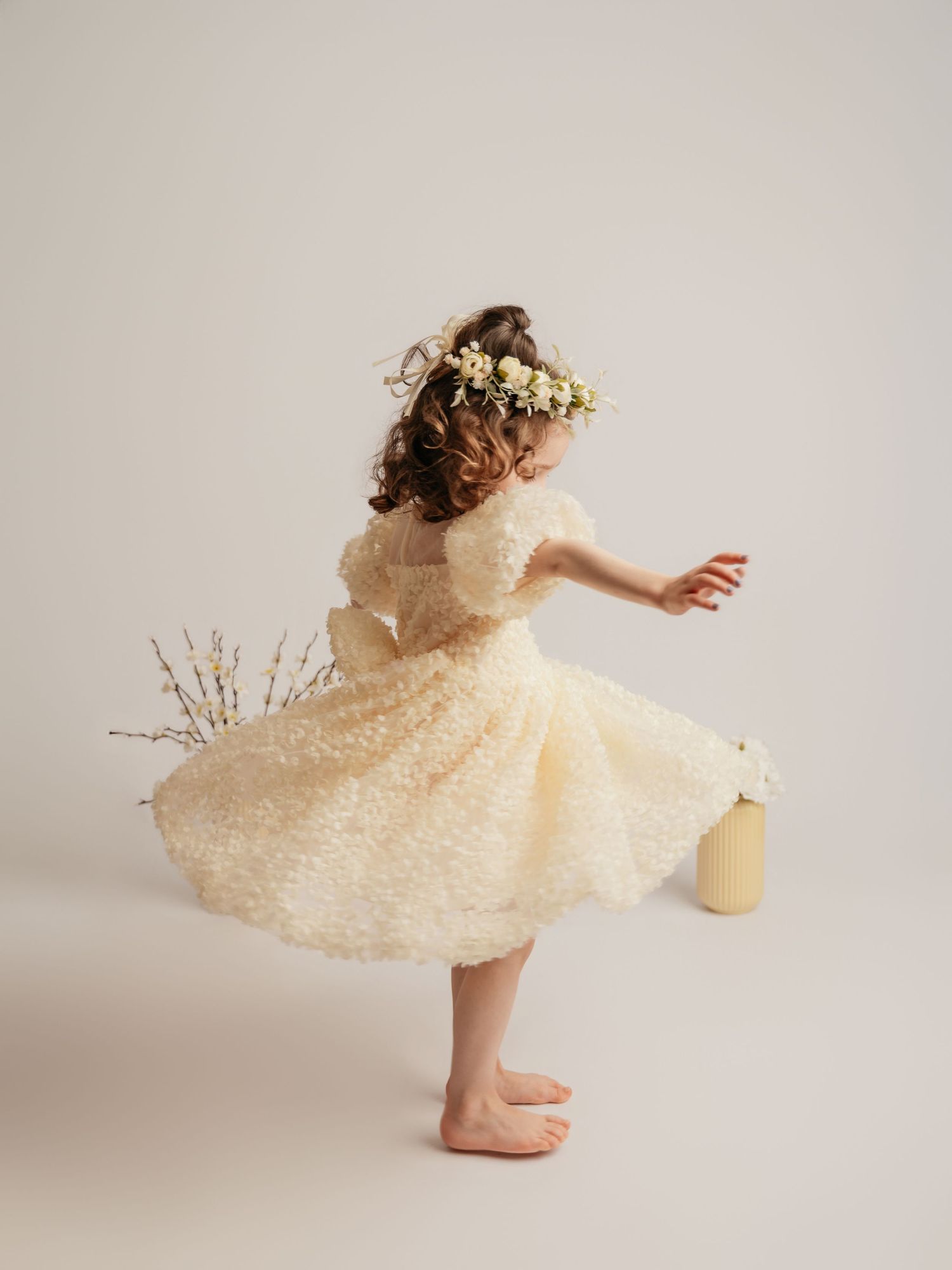 A young girl in a cream textured dress and floral crown twirls barefoot in a bright studio.