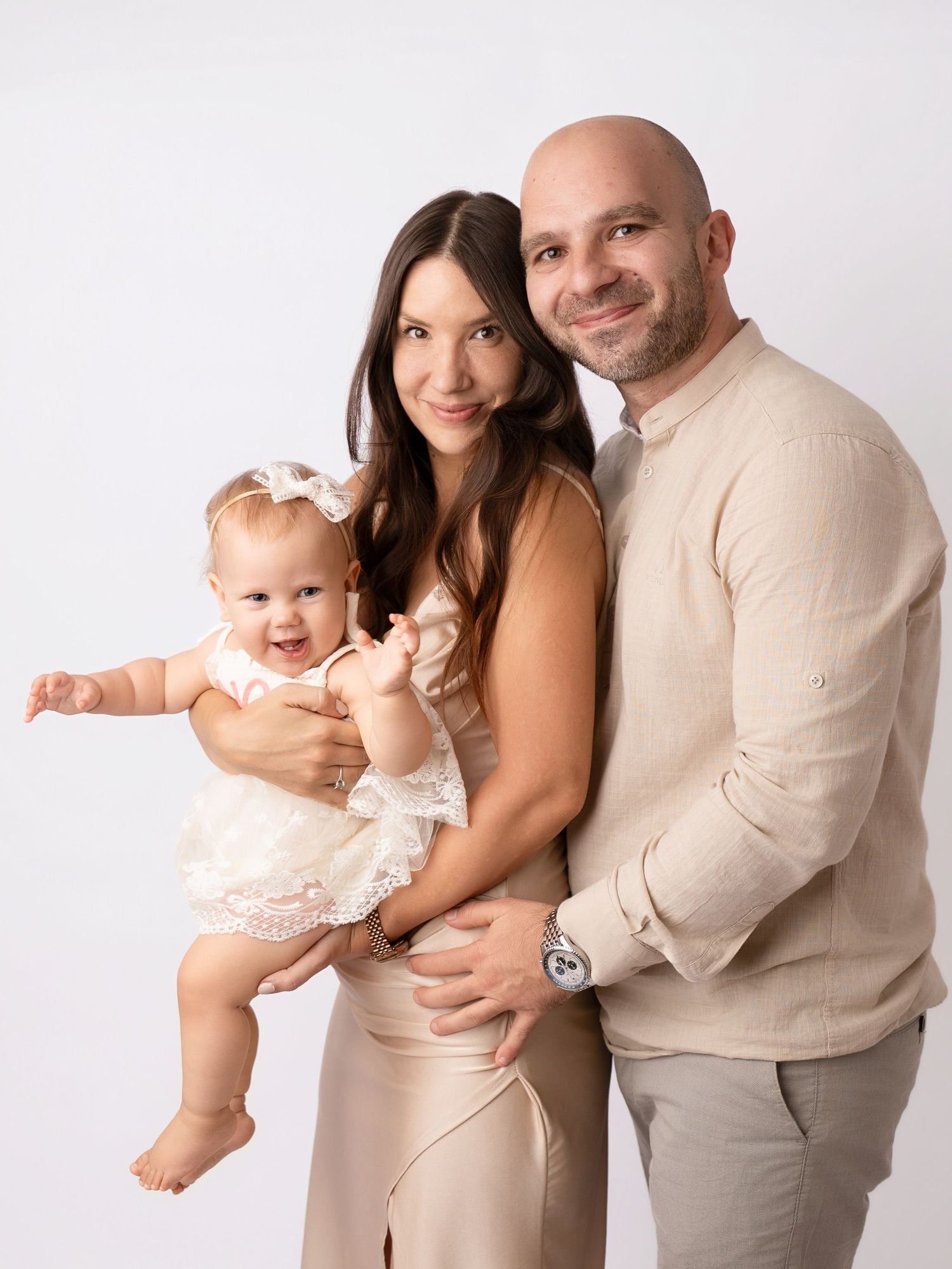 Happy family portrait with parents holding smiling baby girl in white dress against white background.