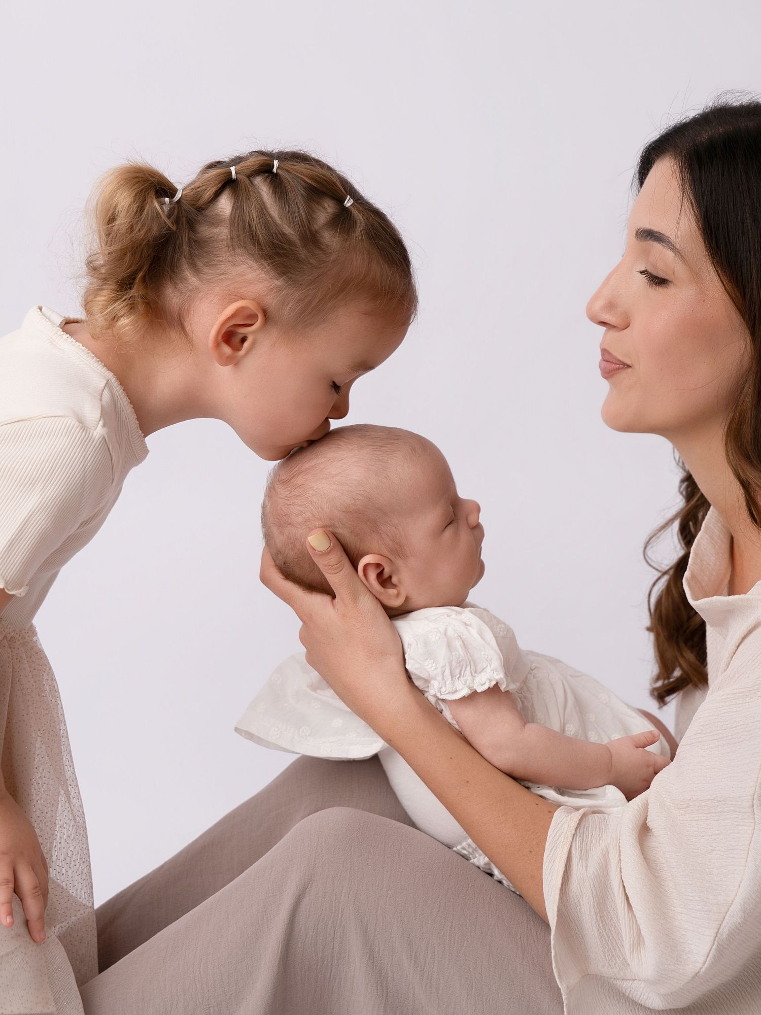 A mother holds her newborn baby while her young blonde daughter leans in to give the infant a gentle kiss.