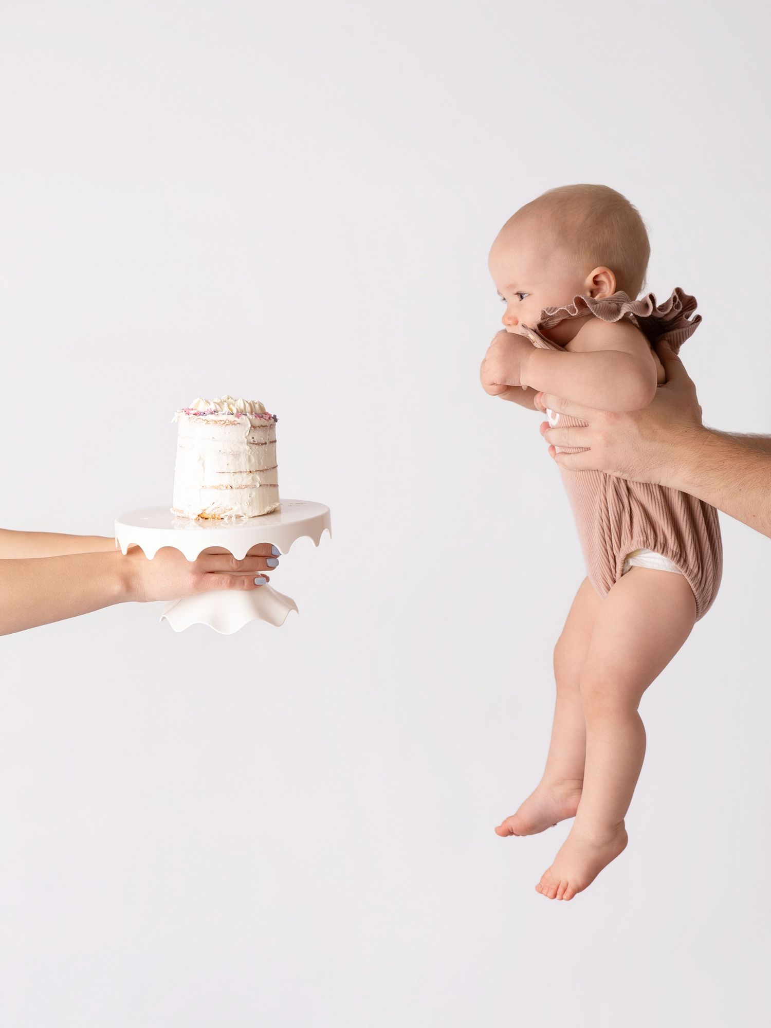 A baby in a brown onesie is held up facing a small birthday cake on a white stand against a light background.
