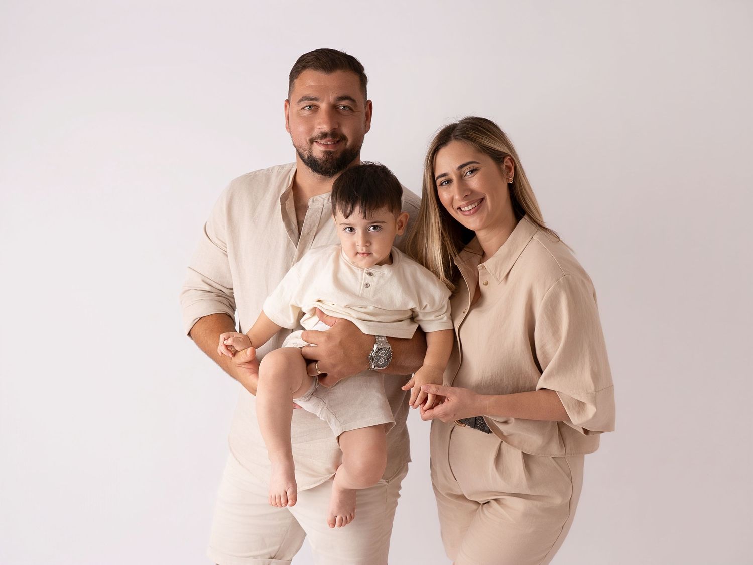 Happy family of three in matching beige outfits pose together in a bright white studio.