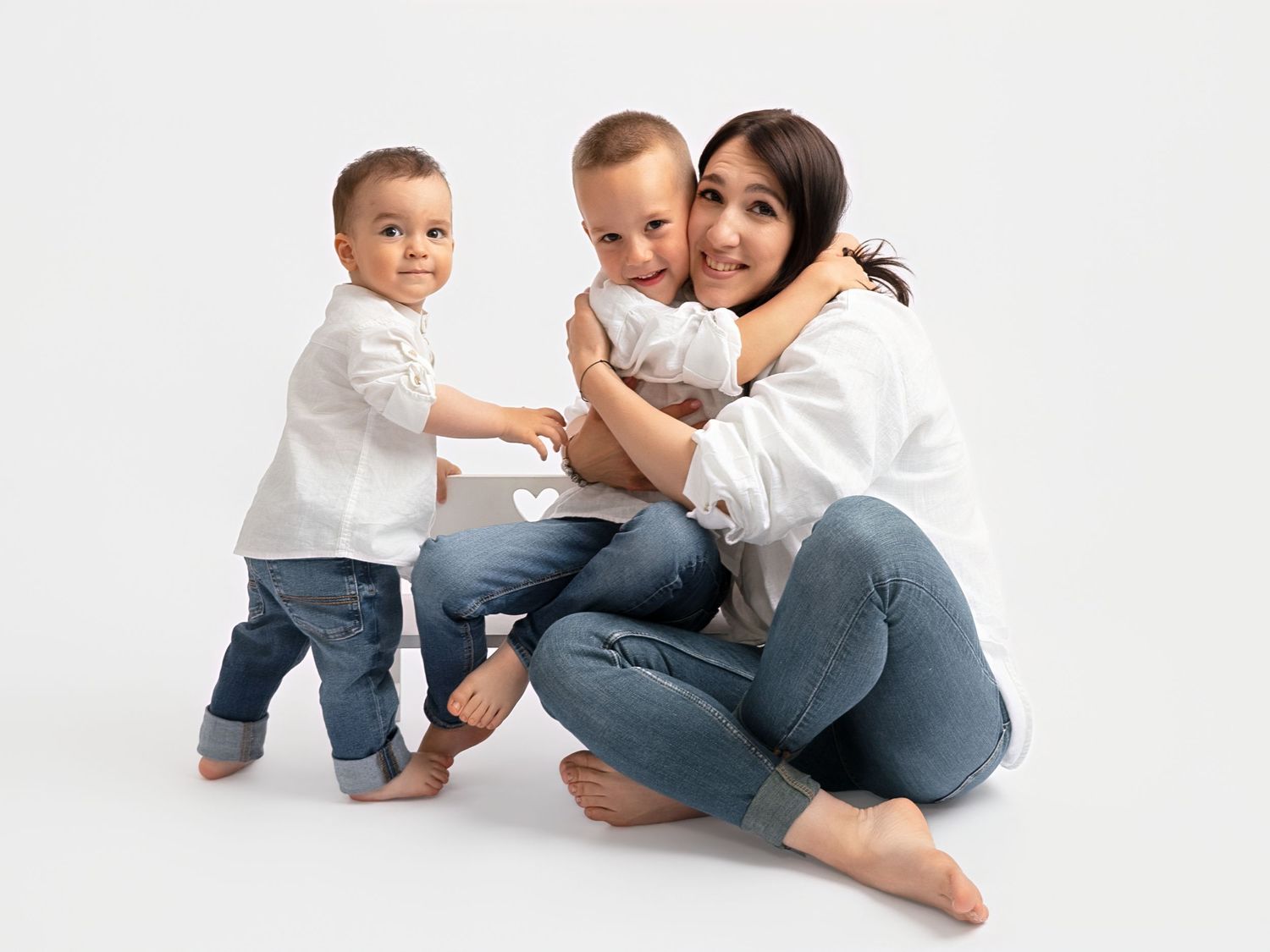 A smiling mother in a white shirt and jeans sits barefoot, hugging two toddlers dressed in matching white shirts and jeans.