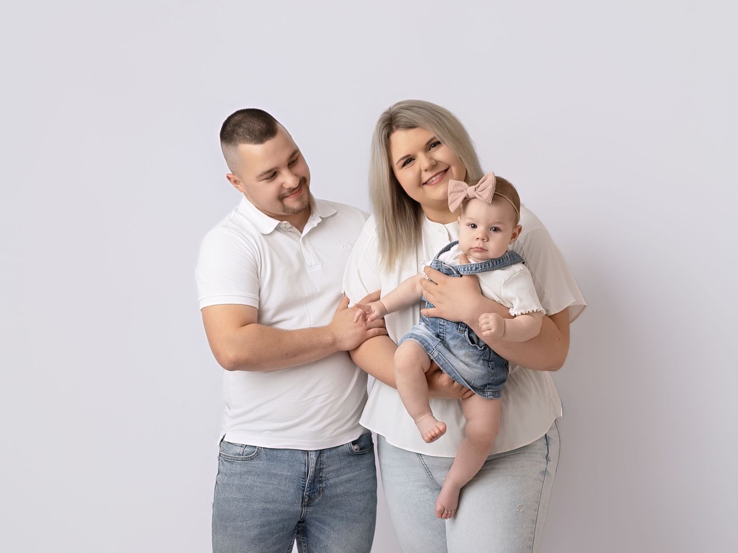A young family posing together, parents in white tops and jeans holding their baby boy against a white background.
