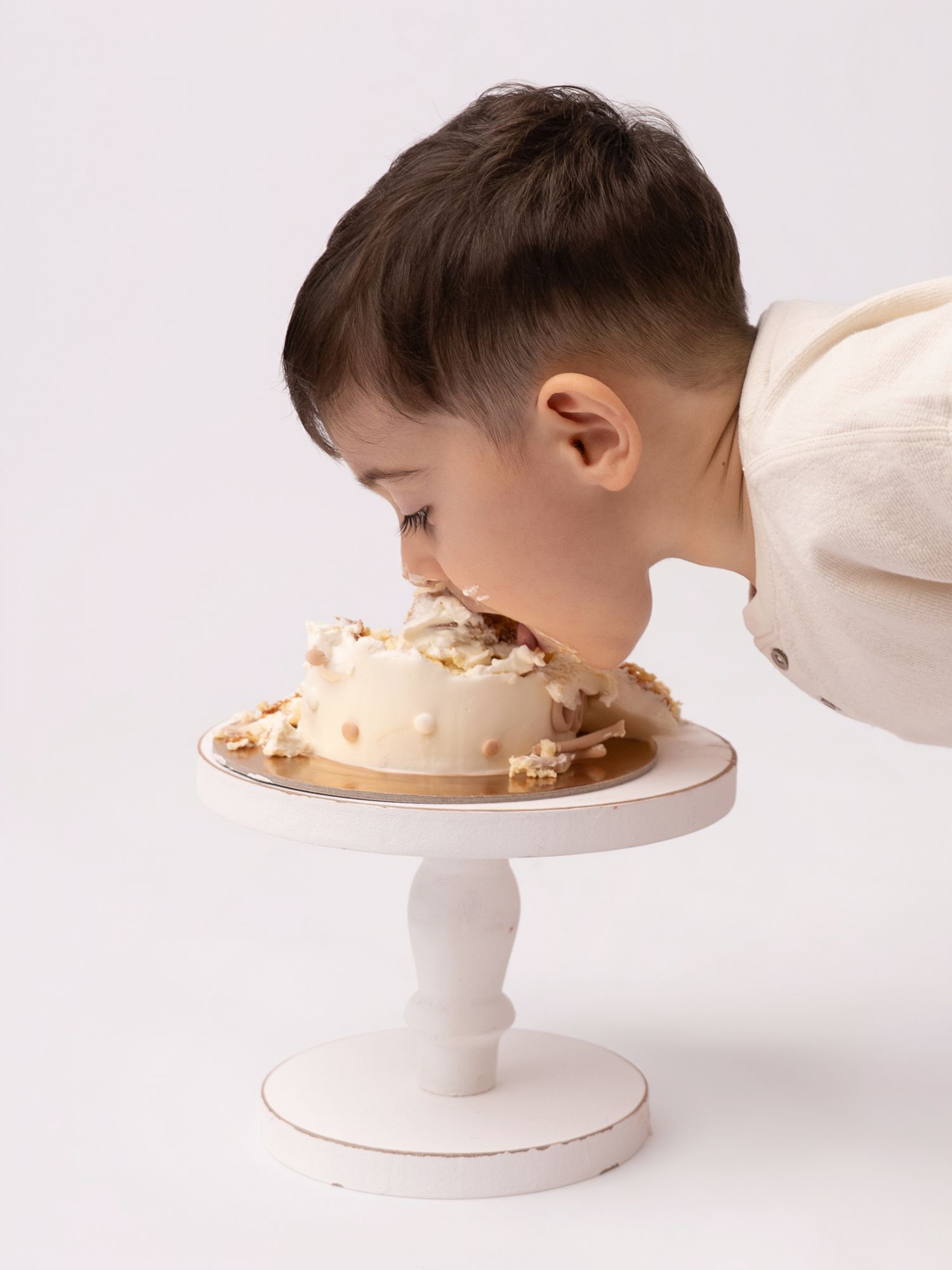 Young boy leaning in to take a bite of a white smash cake on a pedestal stand.